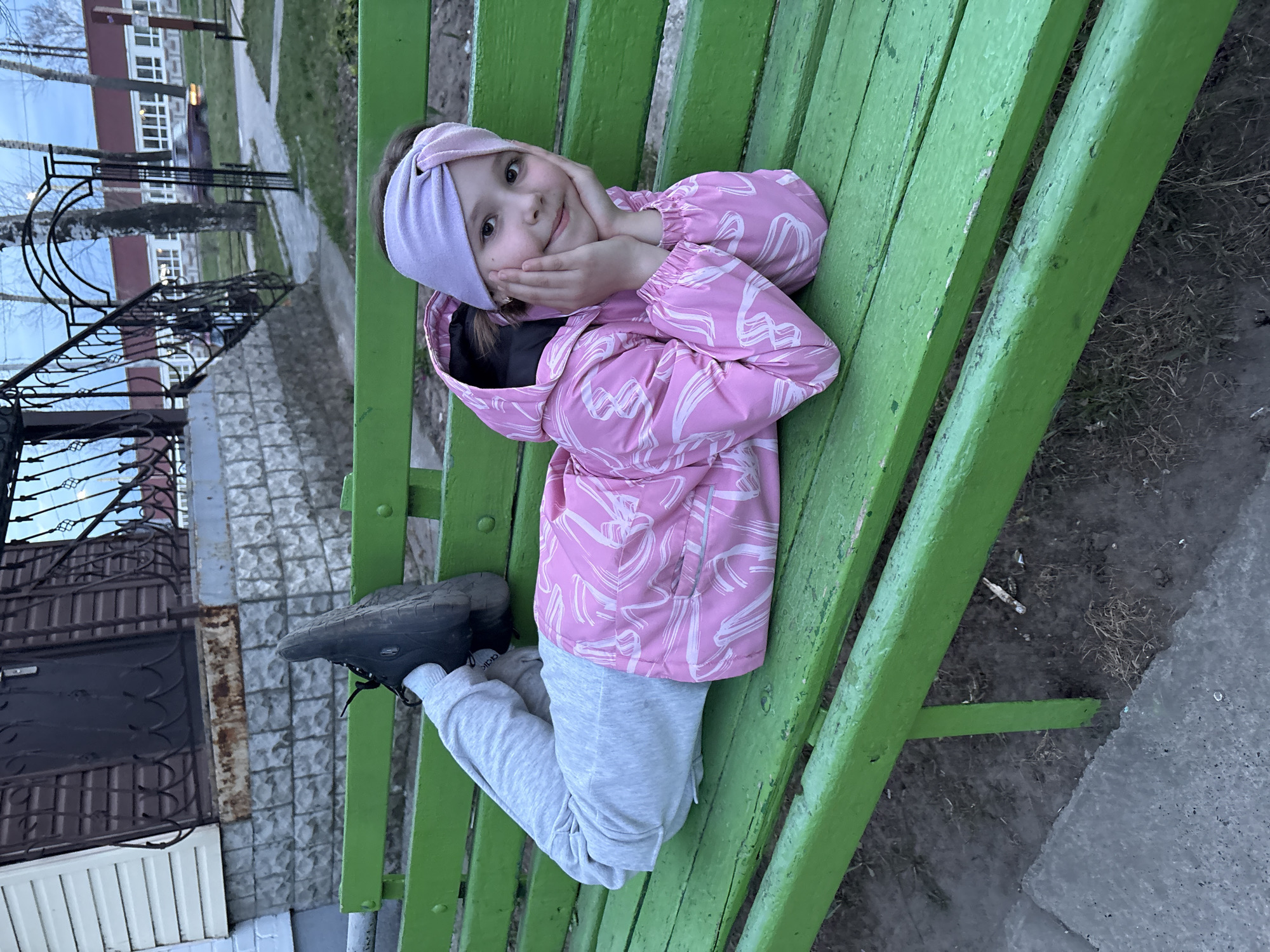 A young girl is lying on a green park bench, facing the camera. She is wearing a pink jacket with white designs, grey sweatpants, a purple headband, and black boots. She is propped up on her elbows, with her hands touching her cheeks. The park bench has a wooden slat design, and the surrounding environment includes a building, a wrought-iron fence, a paved walkway, and trees. Based on previous context, the location is the city of Chaikovsky, Russia.