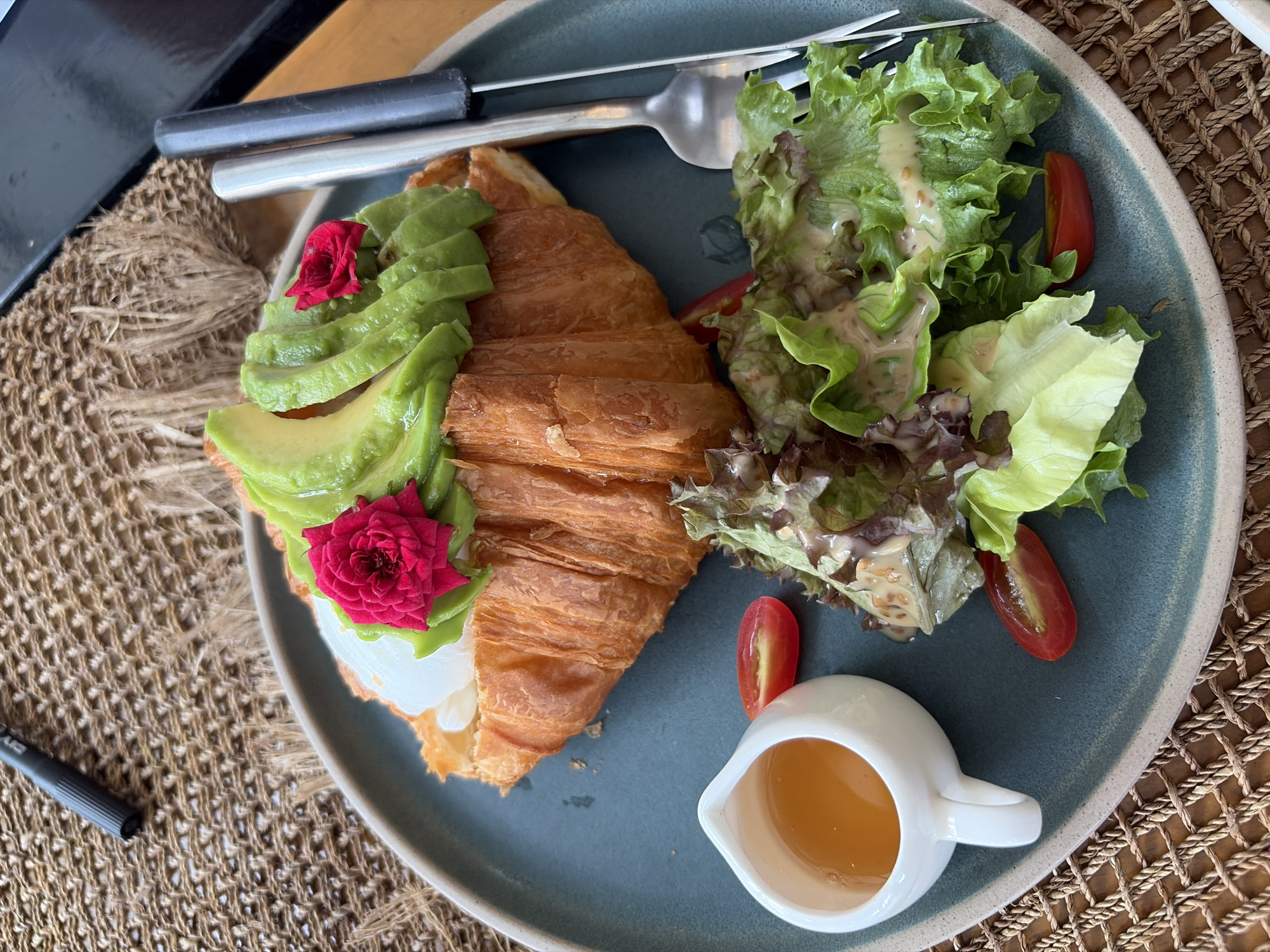 A close-up, overhead shot captures a breakfast dish served on a teal plate, situated on a woven placemat. The plate holds a croissant topped with sliced avocado and two small pink flowers. To the side of the croissant is a small white pitcher filled with golden liquid, likely honey or syrup, and a side salad with leafy greens and cherry tomato halves. A fork and knife are also visible on the right side of the plate. The background is blurred, but hints of a dark surface and a pen suggest a cafe or restaurant setting. The overall scene suggests a meal in Baan Bo Phut, Thailand, likely enjoyed in a relaxed atmosphere.