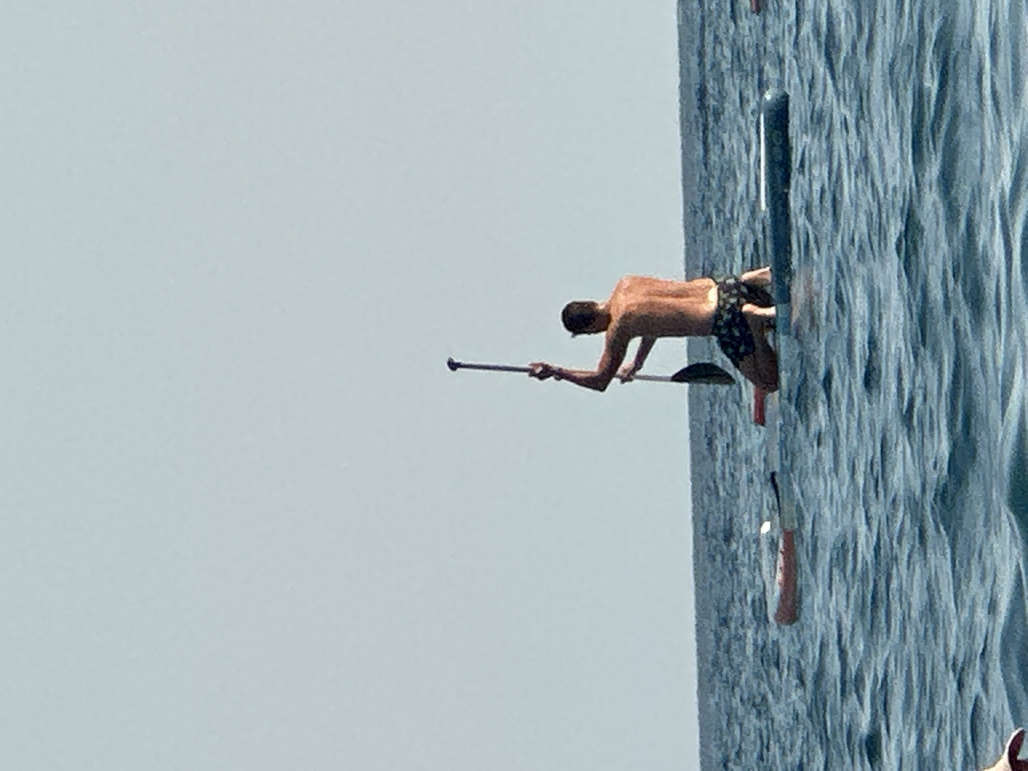 A person is kneeling on a paddleboard in the sea, holding a paddle upright. The person is shirtless and wearing patterned swim trunks. The paddleboard is blue and appears to be floating on calm, blue water. In the background, there is another paddleboard with a red and white object on it. The sky is clear and light blue. This image was captured in Boreti, Montenegro.