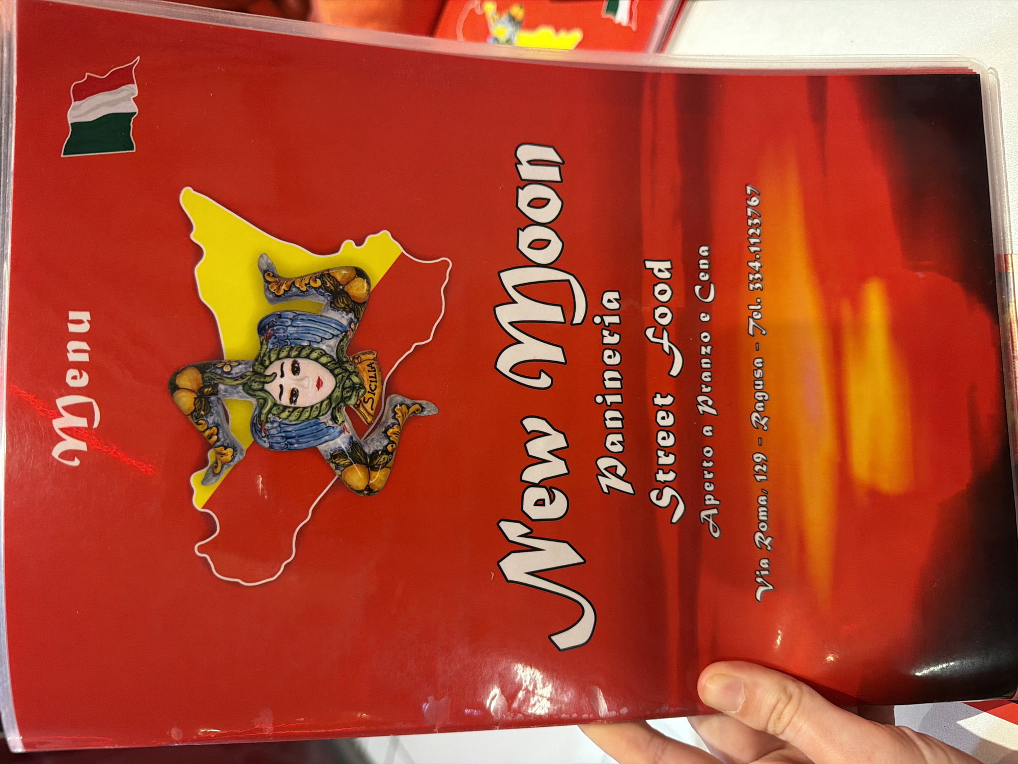 The image shows the menu of a restaurant called "New Moon Panineria Street Food" located at Via Roma, 129 in Ragusa, Italy. The menu is red with the name "New Moon" in large white letters. The restaurant offers street food and is open for lunch and dinner. The menu also features a symbol of Sicily, with the "Trinacria" in yellow outline on a red map background, as well as a Hungarian flag in the upper left corner. The telephone number is 334.1123767. A hand is holding the menu.