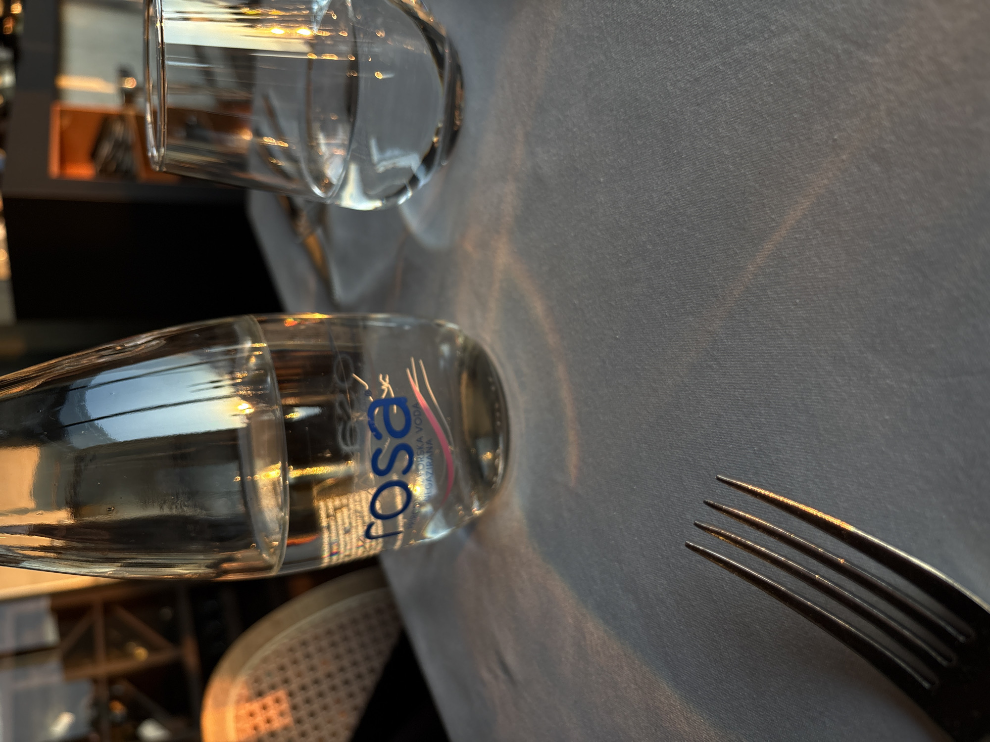 The image depicts a restaurant table setting in Belgrade, Serbia. The tablecloth is light gray. A bottle of "Rosa" brand mineral water lies horizontally on the table. A drinking glass stands above the bottle. To the right, the tines of a fork are visible. The scene suggests a meal setting in a restaurant, with the "Rosa" water bottle providing a clue to the location within Serbia, as it is a locally sourced brand.