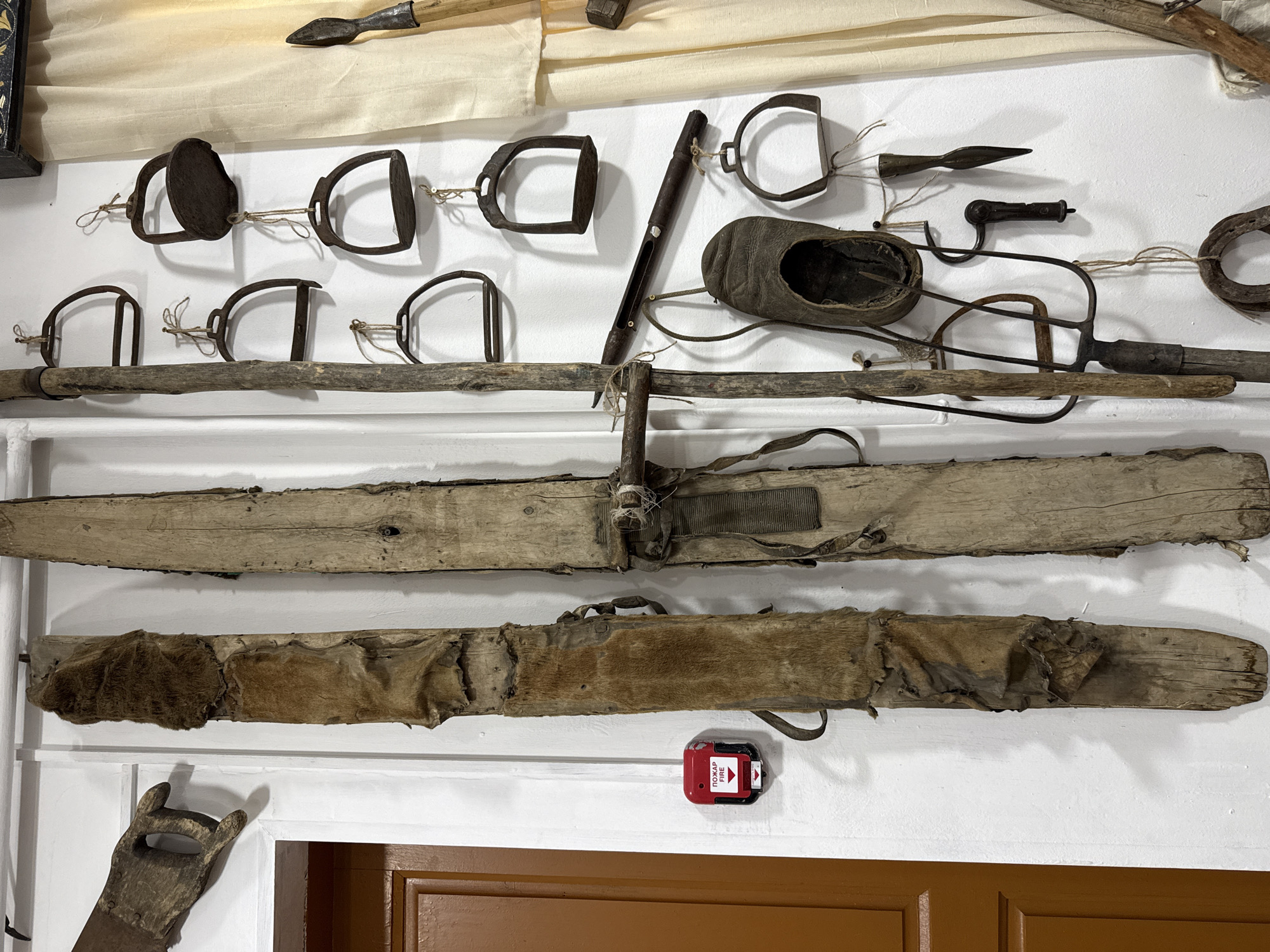 The image presents an indoor display of historical artifacts, possibly within a museum or cultural center, located in Ust-Koksa, Russia. The artifacts are mounted on a white wall above a dark brown door frame.
The artifacts include:
* A collection of metal stirrups and metal tools or implements.
* A worn leather shoe.
* Several lengths of rough-hewn wooden beams, some with tattered leather and fur attached.
* One wooden tool or implement with a handle shaped to resemble a hand.
* A fire alarm pull station affixed to the wall.
The artifacts suggest a focus on rural life, including farming or animal husbandry practices. The overall impression is of an exhibition of ethnographic or historical significance.