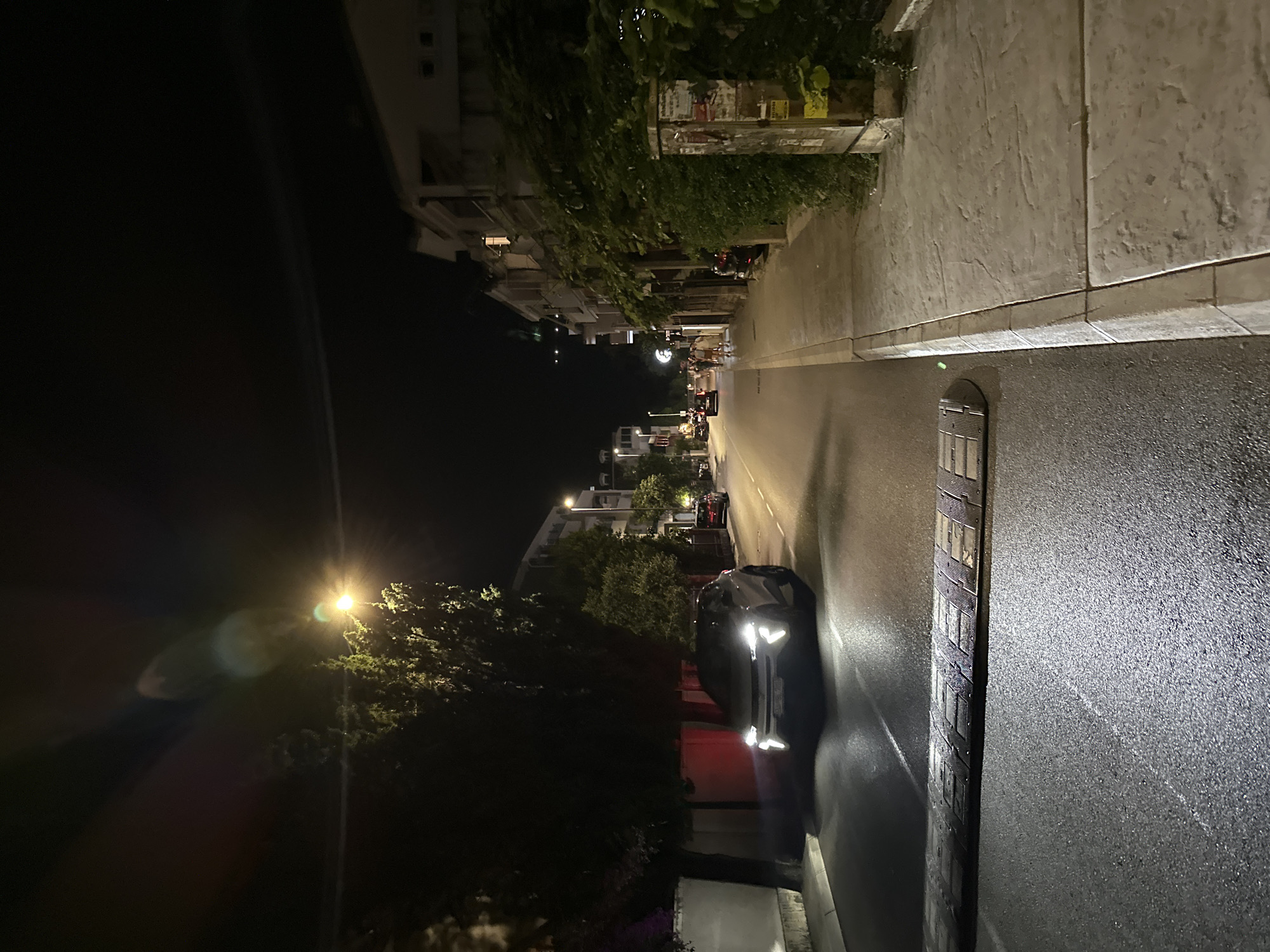 The image depicts a street scene at night in Budva, Montenegro. A white car with its headlights on is parked on the left side of the road, facing away from the camera. In front of the car is a speed bump. The road is wet, reflecting the light from the car and streetlights. Trees line the left side of the street, and buildings with illuminated windows are visible on the right. Several people are walking on the sidewalk on the right side of the street. The sky is dark, indicating it is nighttime.