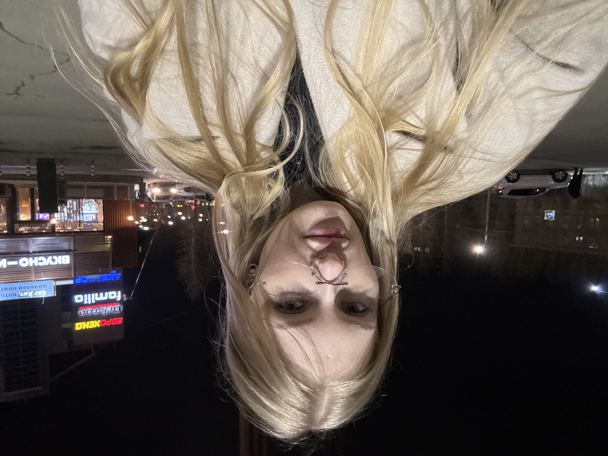 A vertically inverted image of a blonde woman wearing eyeglasses, with street signs visible in the background identifying the location as Perm, Russia. The signs, written in Cyrillic, include "ЕвроХим" (EuroChem), "Familia", and "Вкусно - и точка" (Vkusno - i tochka). A white vehicle is also visible in the background. It is nighttime.