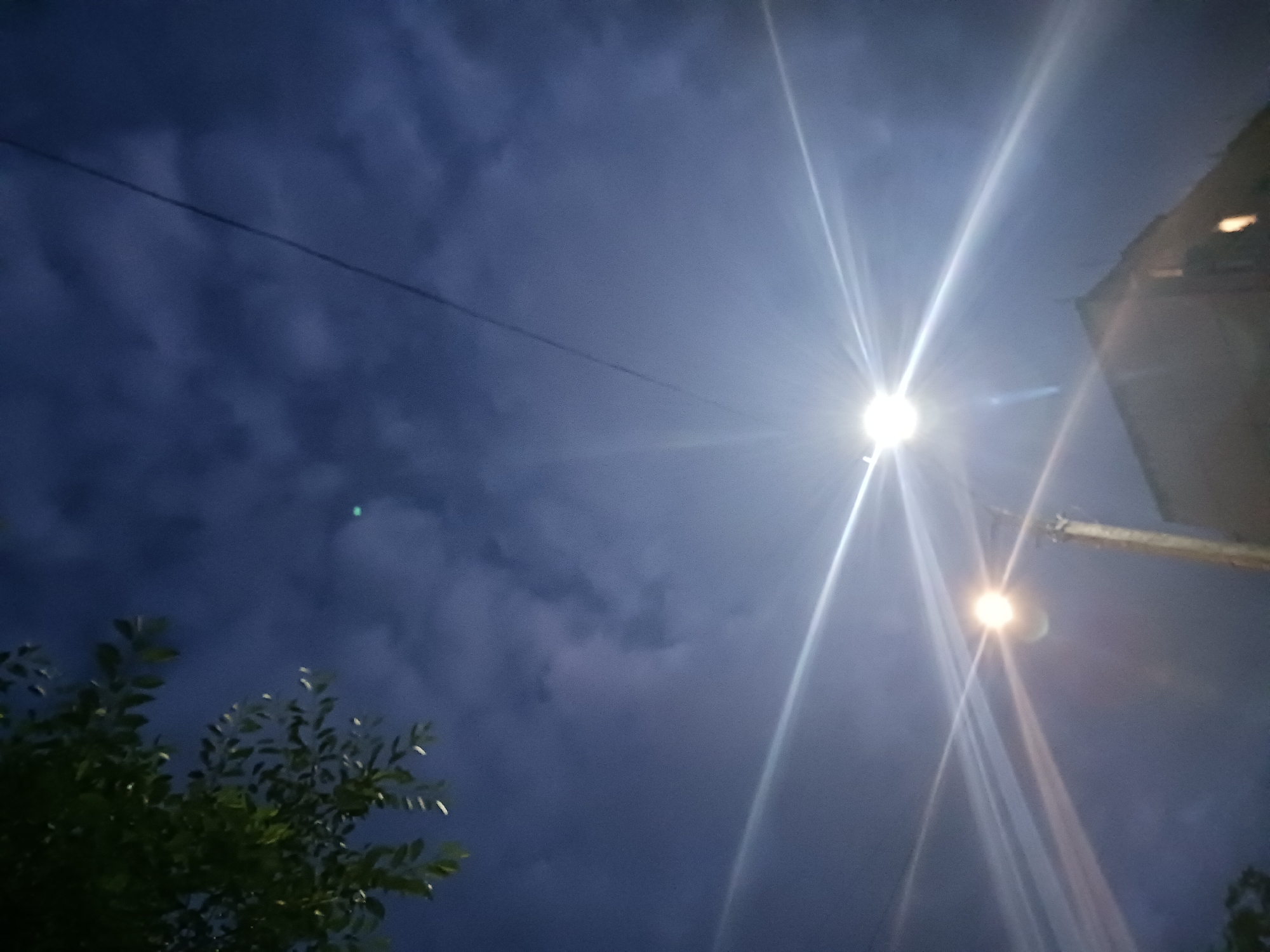 A nighttime, low-angle shot captures a dark blue sky filled with scattered clouds. A streetlamp with a bright, starburst effect is prominently featured in the center, with its light illuminating the surrounding sky and a nearby building. To the left, a dark green tree branch with leaves hangs into the frame. A thin black wire stretches diagonally across the upper portion of the image. The scene appears to be in an urban environment.