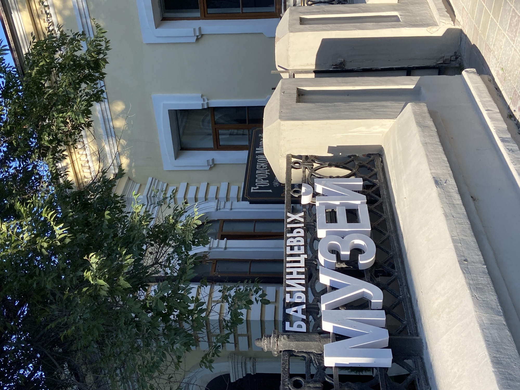 This is a medium shot, eye-level, exterior photograph taken in Blagoveshchensk, Russia. The focal point of the image is a white, three-dimensional sign spelling out "МУЗЕЙ" (Museum) in large, bold letters, mounted on a black metal fence. Above and slightly to the left of this sign, white letters on a black background spell out "БАБИНЦЕВЫХ" (Babintsev's). A smaller black sign with white, ornate Cyrillic lettering is visible behind the "БАБИНЦЕВЫХ" sign, reading "ГОРОДСКОЙ МУ" (City Museum). The sign is attached to a black metal fence with decorative scrollwork. In the foreground, a low, light-colored concrete wall runs horizontally across the bottom of the frame, casting a shadow from the fence onto its surface. To the right of the sign, two light-colored concrete pillars are visible, flanking an opening. The background shows the facade of a building with pale yellow walls, featuring white trim around rectangular windows with dark frames. A large tree with green leaves partially obscures the building and the sky. The lighting suggests a bright, sunny day, with strong shadows. The overall scene depicts the entrance to a museum.