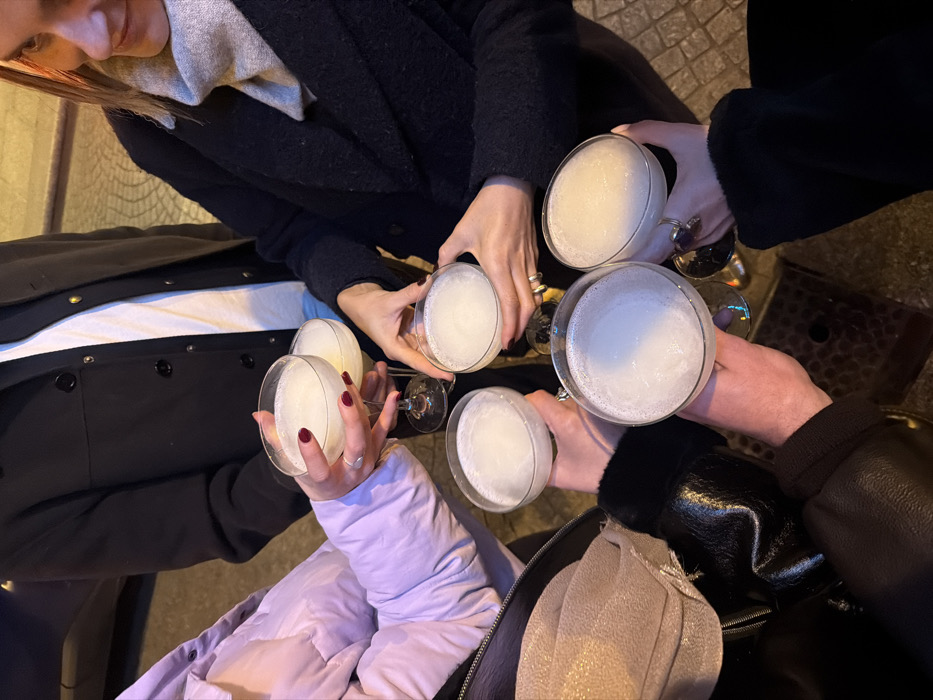 Five individuals clink coupe glasses filled with a creamy, white drink. Their hands, some adorned with rings and manicured nails, hold the stems. The shot is taken from above, capturing the group in their outerwear. A black coat, a lavender puffer, and a leather jacket with a scarf are visible. A patterned bag sits near one of the drinkers.