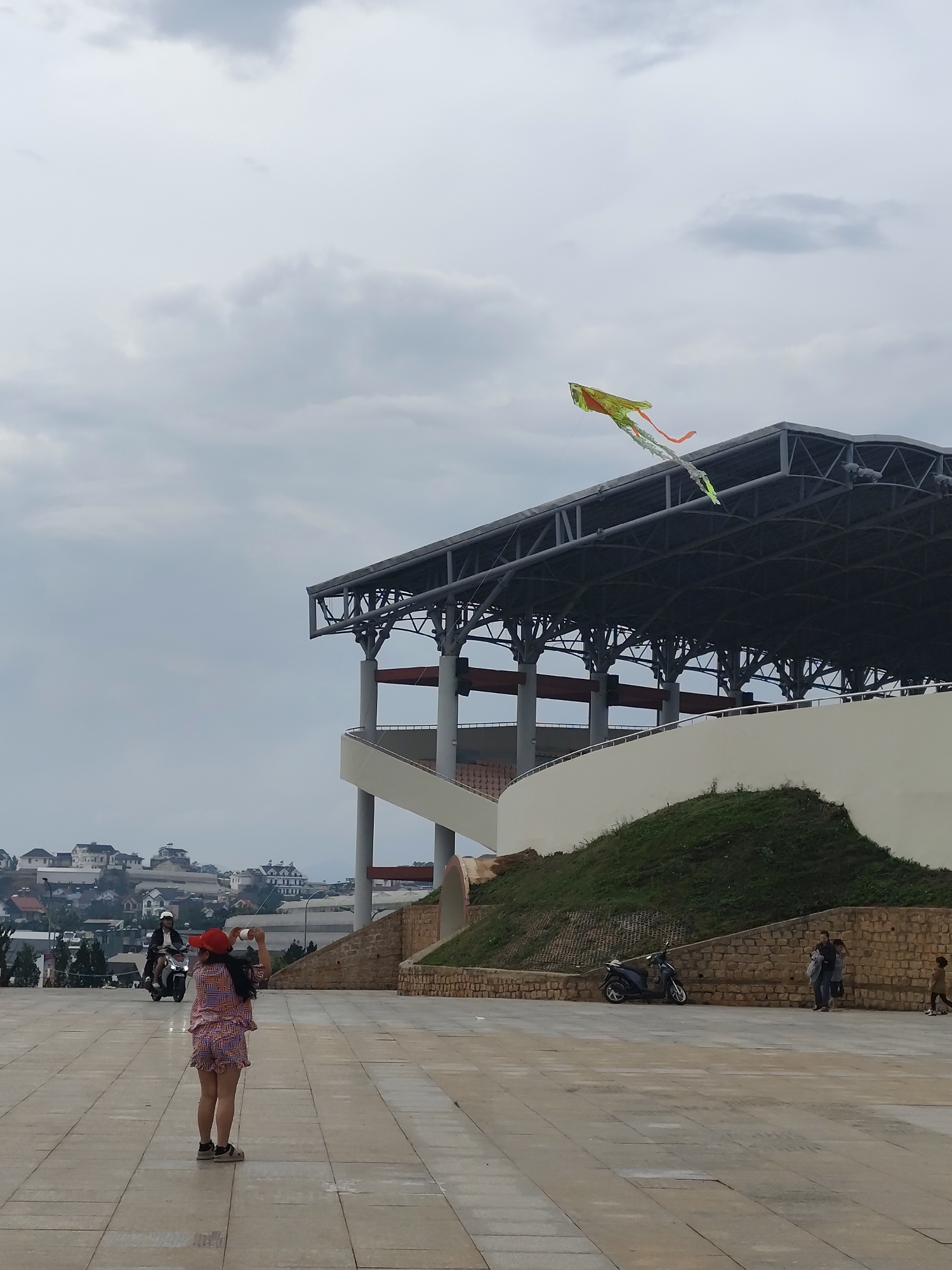 A full shot taken during the daytime shows a public square in the city of Lang Biang - Da Lat, Vietnam. A girl in patterned shorts and a matching shirt and red cap is facing away from the camera, appearing to take a photo. A yellow and orange kite flies above her. A building with a metal frame and a curved staircase is behind her to the right. In the background, behind the girl, a motorcyclist is riding uphill toward the camera. A smaller hill is to the right of the girl and a few pedestrians are near it. A motorbike is parked by the hill. The sky is cloudy.