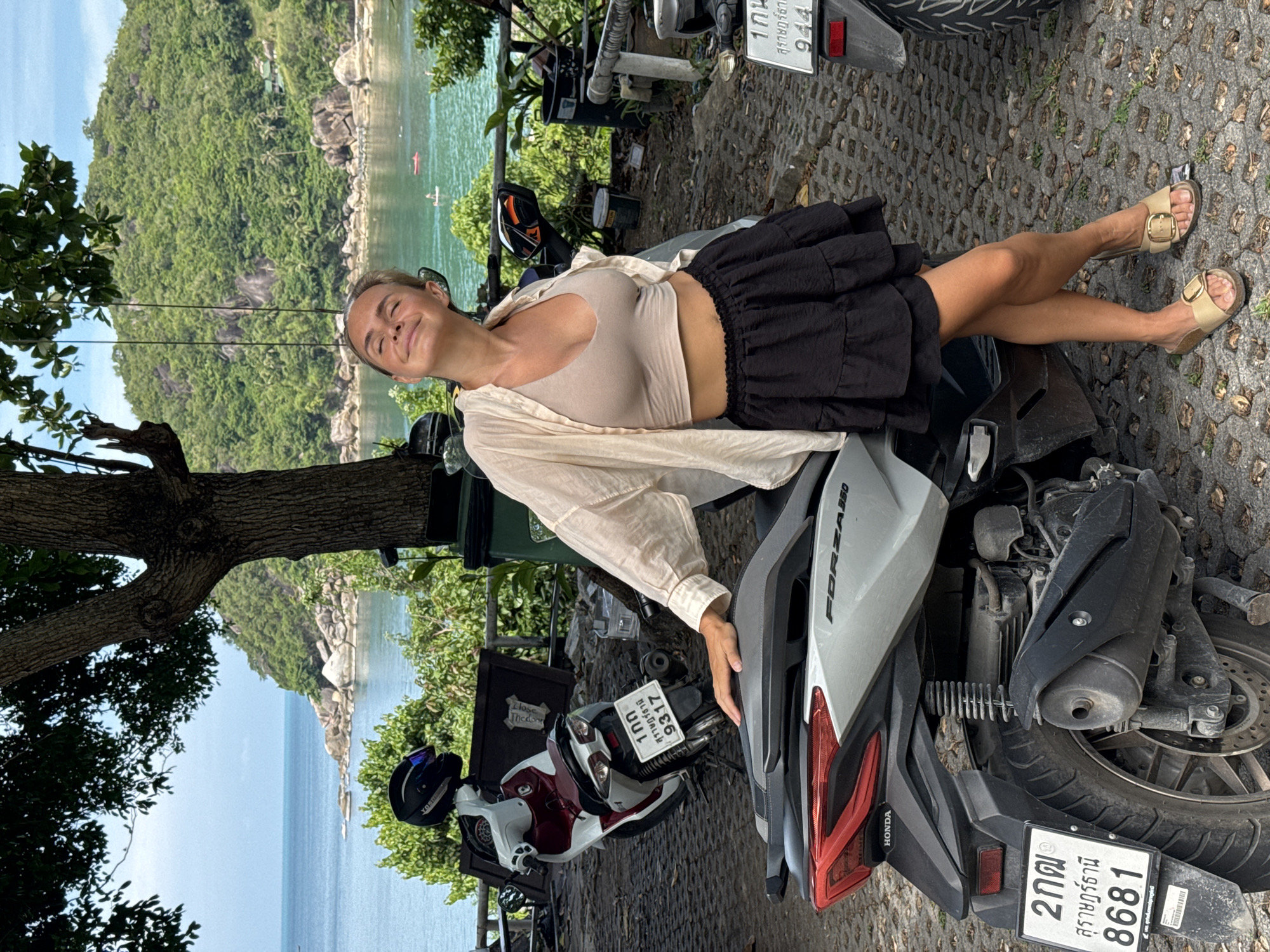 A young Caucasian woman with blonde hair, wearing a beige crop top and a black ruffled skirt, stands on a paved surface next to a silver Honda Forza scooter. She has her eyes closed and her head tilted back, appearing to enjoy the sunshine. A light-colored open shirt is draped over her shoulders. In the background, a lush green hillside overlooks a calm blue ocean with a few boats on it. To the left of the woman, another scooter, white and red, is parked with a black helmet on its seat. The location appears to be a scenic viewpoint on the island of Ko Samui, Thailand.