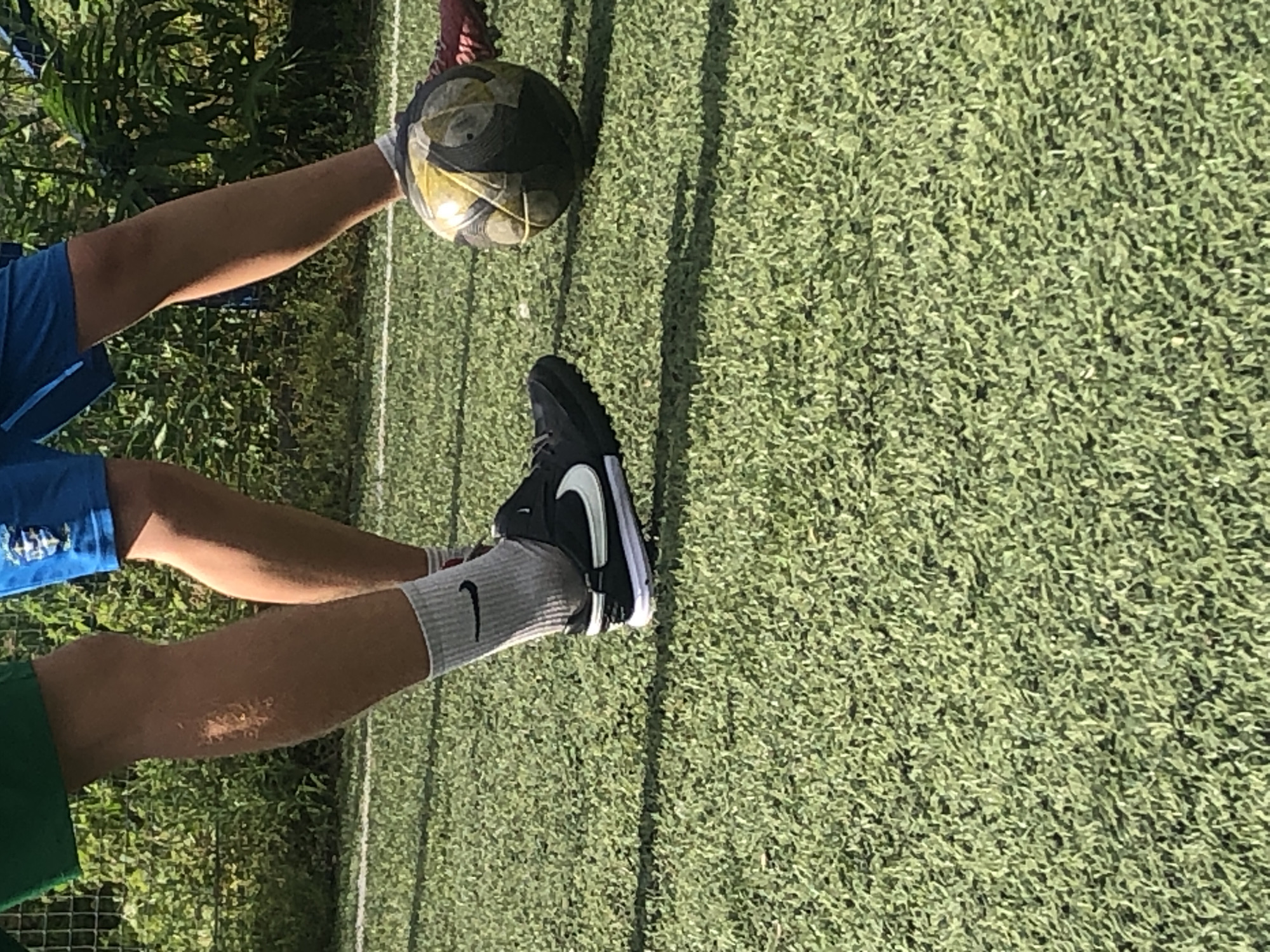 Two individuals are playing soccer on an artificial turf field. One individual is wearing green shorts and a gray sock, with a black Nike soccer cleat on their left foot. The other individual is wearing blue shorts and a white sock. A soccer ball, with yellow and black panels, is visible on the field, being kicked by the individual in blue shorts. The background consists of green foliage and a fence. The image does not provide any information about the specific location within the city of Dnipro, Ukraine.