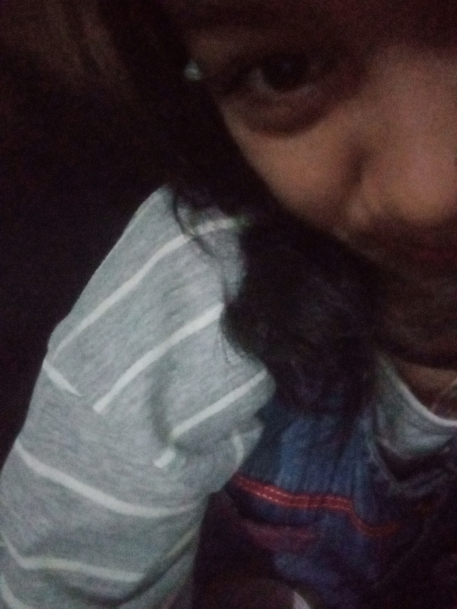 The image is a close-up, low-light selfie of a person's face and upper body, taken indoors. The subject's face is partially obscured by dark hair, and only one eye is clearly visible. They are wearing a grey and white striped long-sleeved shirt, with a glimpse of a dark blue garment with a red stripe underneath. The background is dark and indistinct. There are no discernible details indicating the location within the city of Badas, Indonesia.