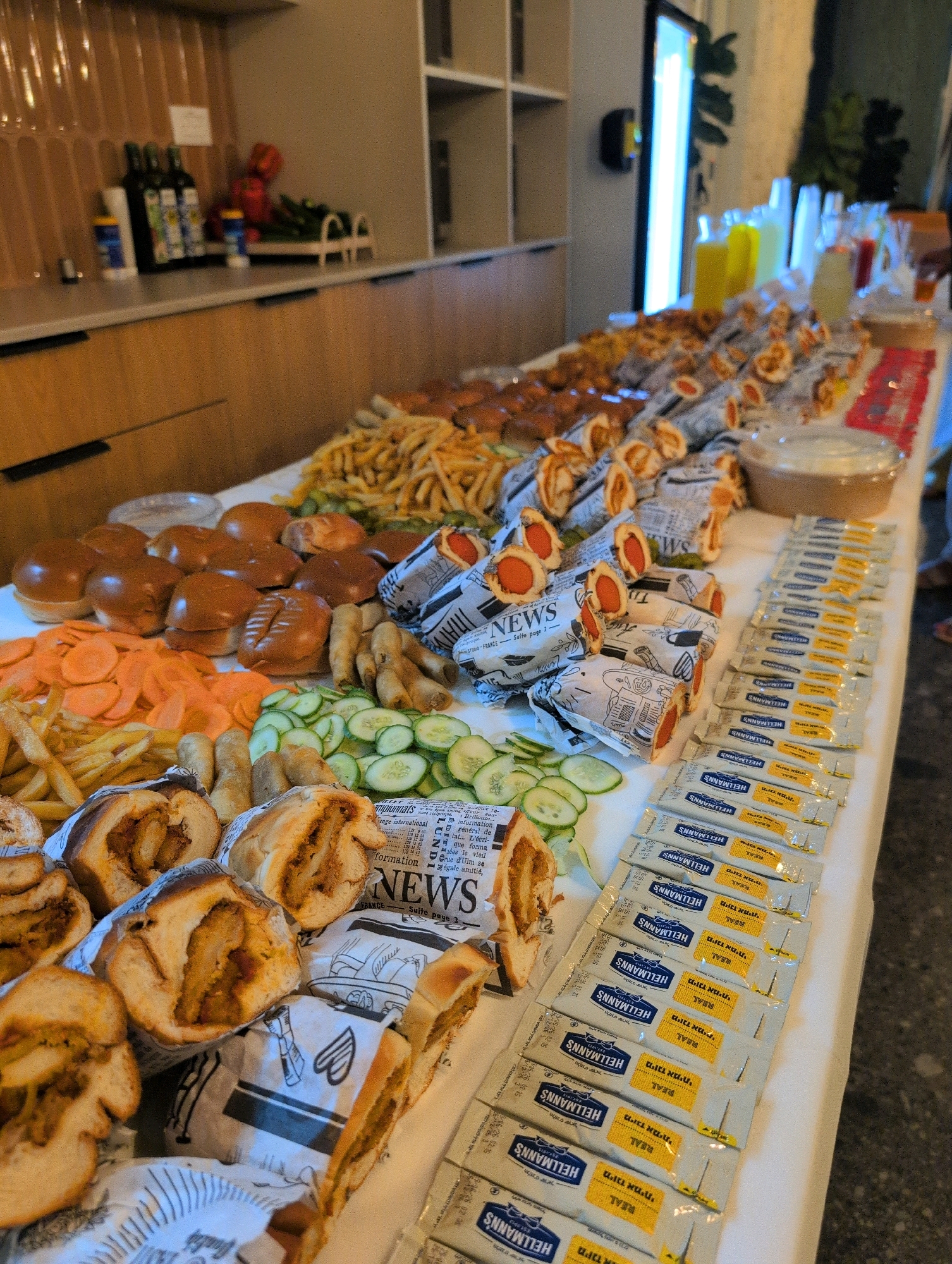 A long buffet table is laden with food, likely for a catering event or a party. The table is covered with a white tablecloth and extends from the left foreground to the right background of the image.
In the foreground on the left, there are several halves of large sandwiches, showing a filling of what appears to be shredded meat and sauce. To the right of these sandwiches are rows of individual condiment packets, predominantly Hellmann's brand, with some packets featuring yellow labels and Hebrew text.
Moving towards the center of the table, there are piles of food items. These include numerous burger buns, sliced carrots, french fries, sliced cucumbers, and several small fried rolls or spring rolls. Further back on the table, there are many hot dogs wrapped in newspaper, with the ends of the hot dogs visible. In the background, there are drinks in clear plastic cups and bottles, and a round container of what might be dip or sauce.
The background of the image shows a kitchen or service area. To the left, there are wooden cabinets with metal handles and a countertop with bottles of olive oil and other items. A wall with textured tiling is visible behind the countertop. To the right and center background, there are shelves and part of a tall illuminated vertical display or sign.
The image quality is somewhat blurry, but the arrangement suggests a self-serve food station. The presence of specific condiment brands and the types of food indicate a casual dining or fast-food inspired spread. The context of Rishon LeZion, Israel, suggests this event is taking place in that city.