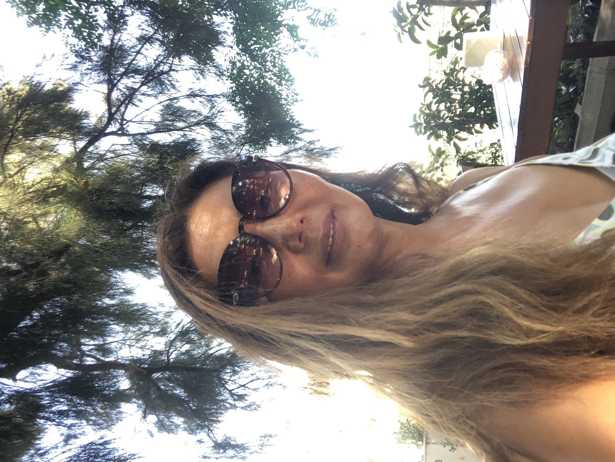 An outdoor, medium-shot, selfie-style photograph depicts a woman with light brown, wavy hair and sunglasses. She is wearing a dress with a pattern of white, grey, and green. The woman is outdoors, with dappled sunlight filtering through the branches of a tree. In the background, there is a table with a white square object and a glass object. Green foliage is visible behind the table. The scene appears to be in the city of Karaburun, Turkey.