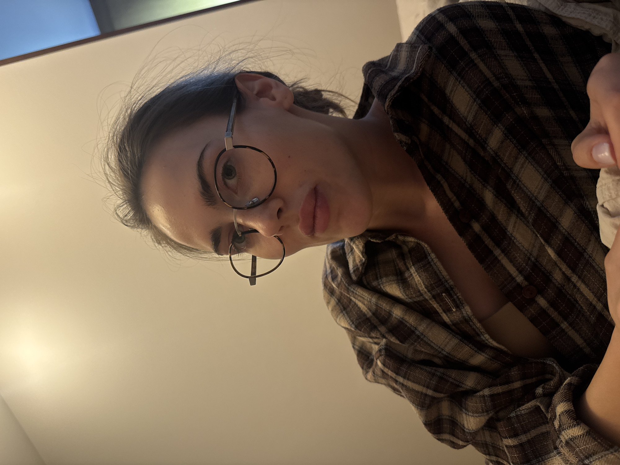 A person with dark hair pulled back, wearing round glasses and a brown and tan plaid shirt, is looking down at the camera. The background is a plain white wall. The location and city are unknown.