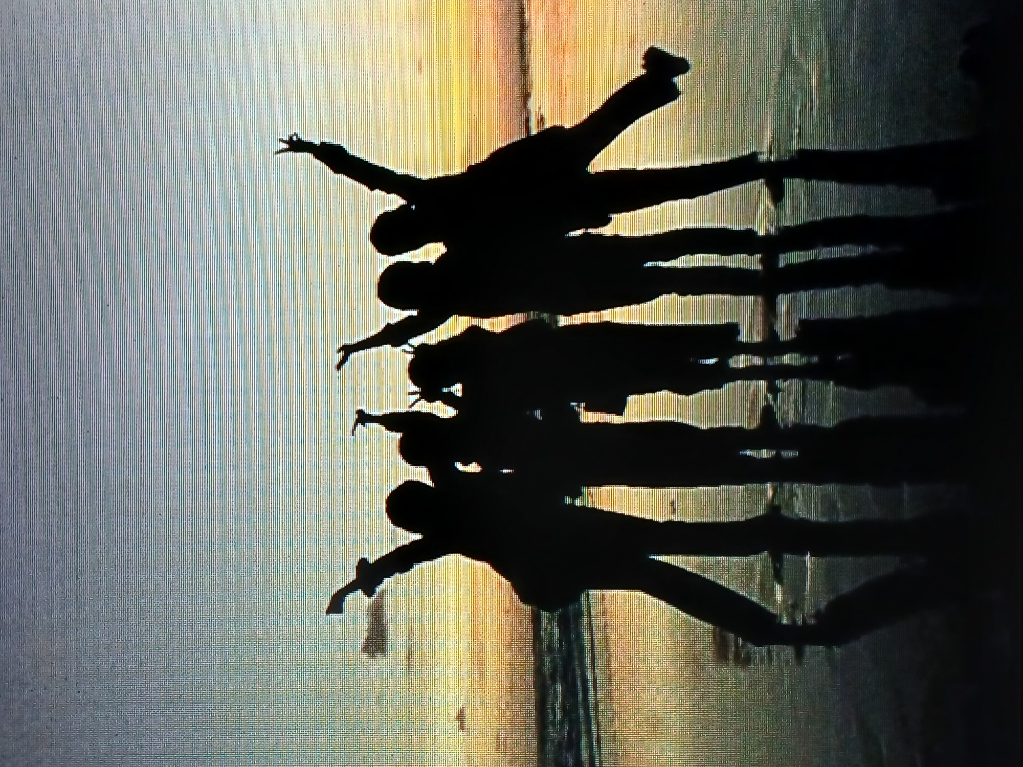 A group of six silhouetted figures are captured against a backdrop of an ocean horizon and sunset, likely in Juan Griego, Venezuela. The figures appear to be standing in a line or formation on what could be a beach or shoreline, casting shadows onto the ground below. Each person is in a different pose, some with arms outstretched or legs raised, suggesting movement or playfulness. The sky is a blend of colors, transitioning from a light hue near the horizon to a darker shade above. The lighting conditions indicate it is either sunrise or sunset, which are common occurrences in Juan Griego due to its coastal location. The image conveys a sense of camaraderie and enjoyment of the coastal environment.