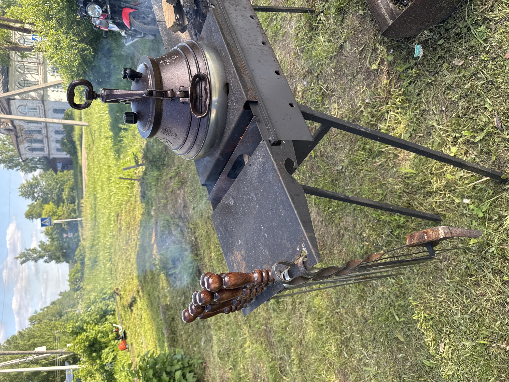 An outdoor scene in Луптюг, Russia, captures a cooking setup under bright daylight. In the foreground, a large, dark-colored, ornate pot with a distinctive handled lid sits atop a black metal brazier or grill stand. Smoke gently rises from beneath the pot, indicating a fire is burning and cooking is in progress. The pot appears to be made of cast iron or a similar heavy material, featuring embossed decorative patterns around its body.
To the right of the pot, an array of metal skewers with polished wooden handles is neatly arranged on a rack, suggesting preparations for grilling alongside the pot-cooked dish. The brazier itself is a sturdy black metal structure with several vents or holes along its sides.
The setting is a grassy area. In the middle ground to the left, a large, weathered multi-story building with multiple arched windows and intricate architectural details stands, hinting at a historical or traditional local structure. Further back, a red and black motorcycle is partially visible, parked on the grass. The background is dominated by lush green trees and foliage under a blue sky with scattered white clouds. A blue road sign with a white arrow is visible in the distance, suggesting proximity to a road. The overall impression is a casual outdoor cooking event, possibly a picnic or a gathering in a rural or semi-urban environment. No people or legible text are visible.