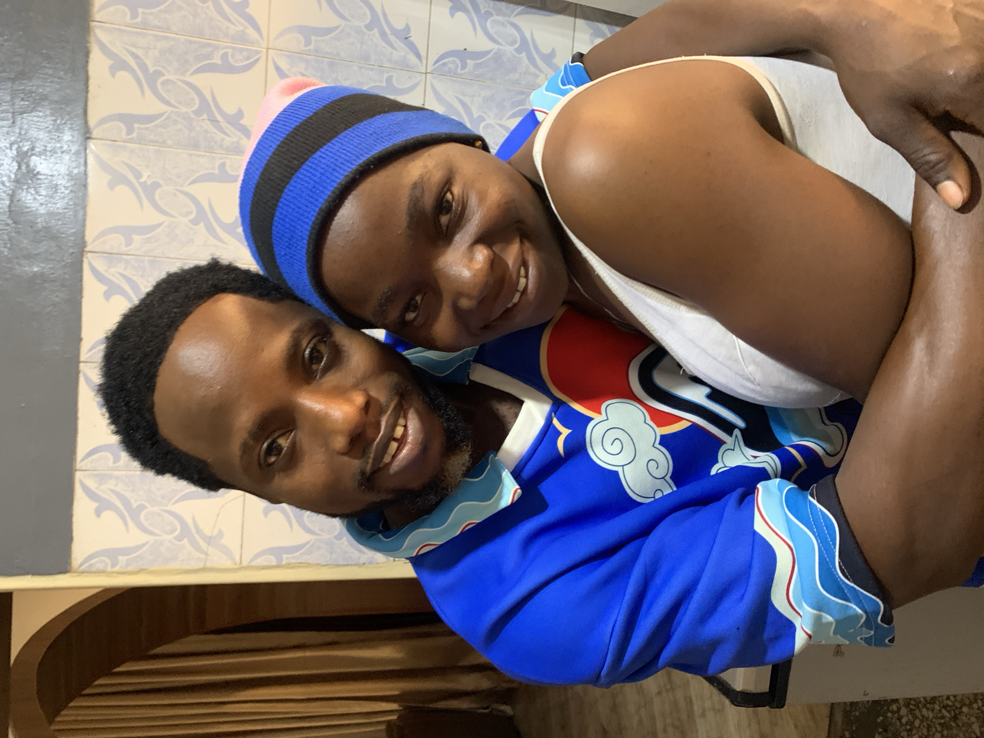 A dark-skinned man and woman are embracing in an indoor setting. The man has a short beard and is wearing a blue jacket with white cloud patterns. The woman is wearing a blue and black striped hat with a pink top, along with a white tank top. They are both smiling. The background includes patterned blue and white tiles. The location is likely a residence in the city of Jos, Nigeria.