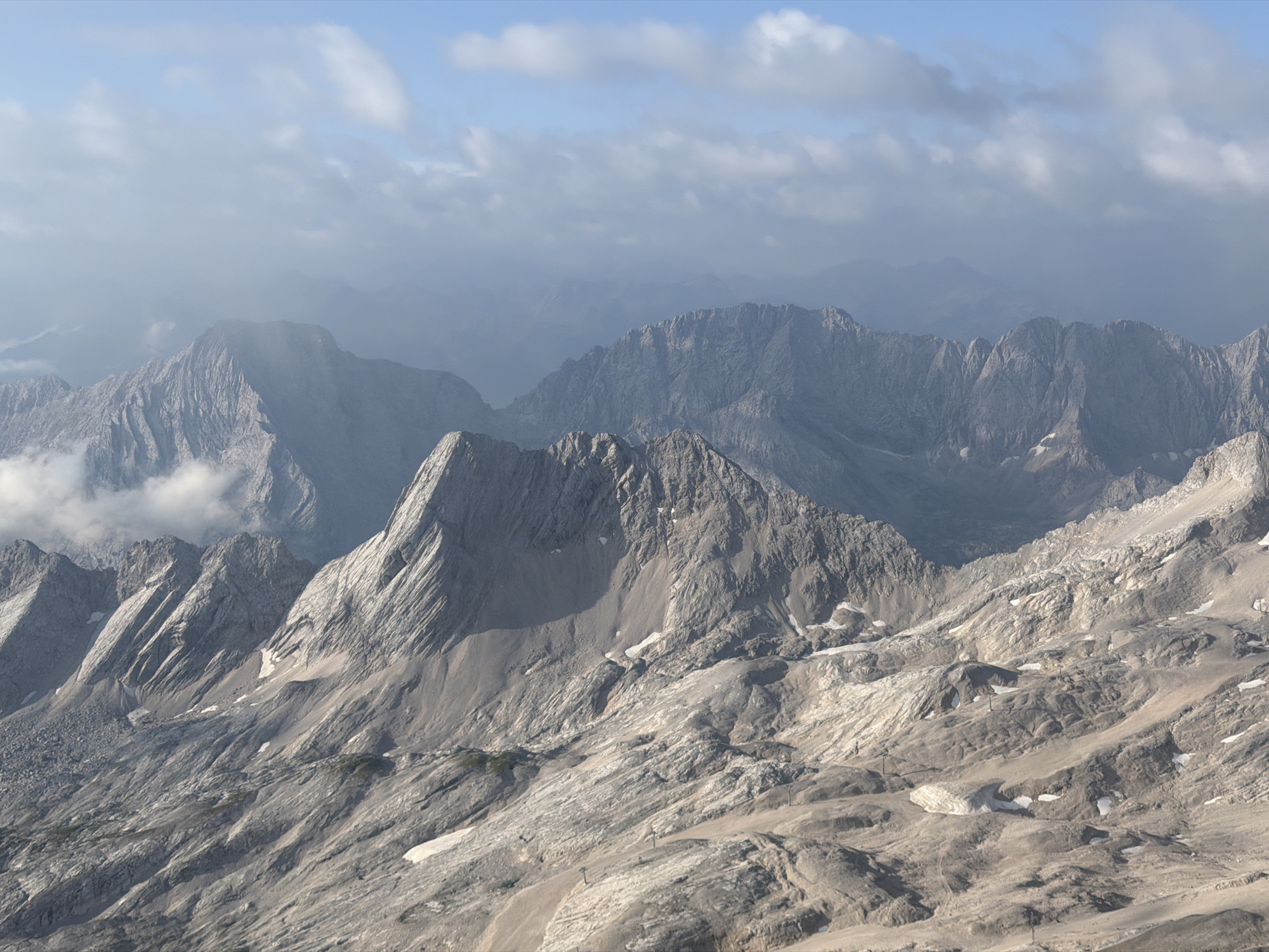 This is an aerial photograph of a mountainous landscape, likely taken from a high vantage point. The scene is dominated by rugged, grey mountain peaks, some of which are partially obscured by clouds and mist. The terrain is rocky and uneven, with visible scree slopes and patches of snow in the shadowed areas. In the foreground, the mountains have a more detailed, textured appearance, with sharp ridges and steep inclines. Further in the distance, the mountains appear softer and more blurred due to atmospheric haze. The sky above is a pale blue, with scattered white clouds. The overall impression is one of a remote and wild alpine environment. This image is identified as being taken in the vicinity of Grainau, Germany, which is known for its proximity to the Bavarian Alps, including the Zugspitze, Germany's highest mountain.