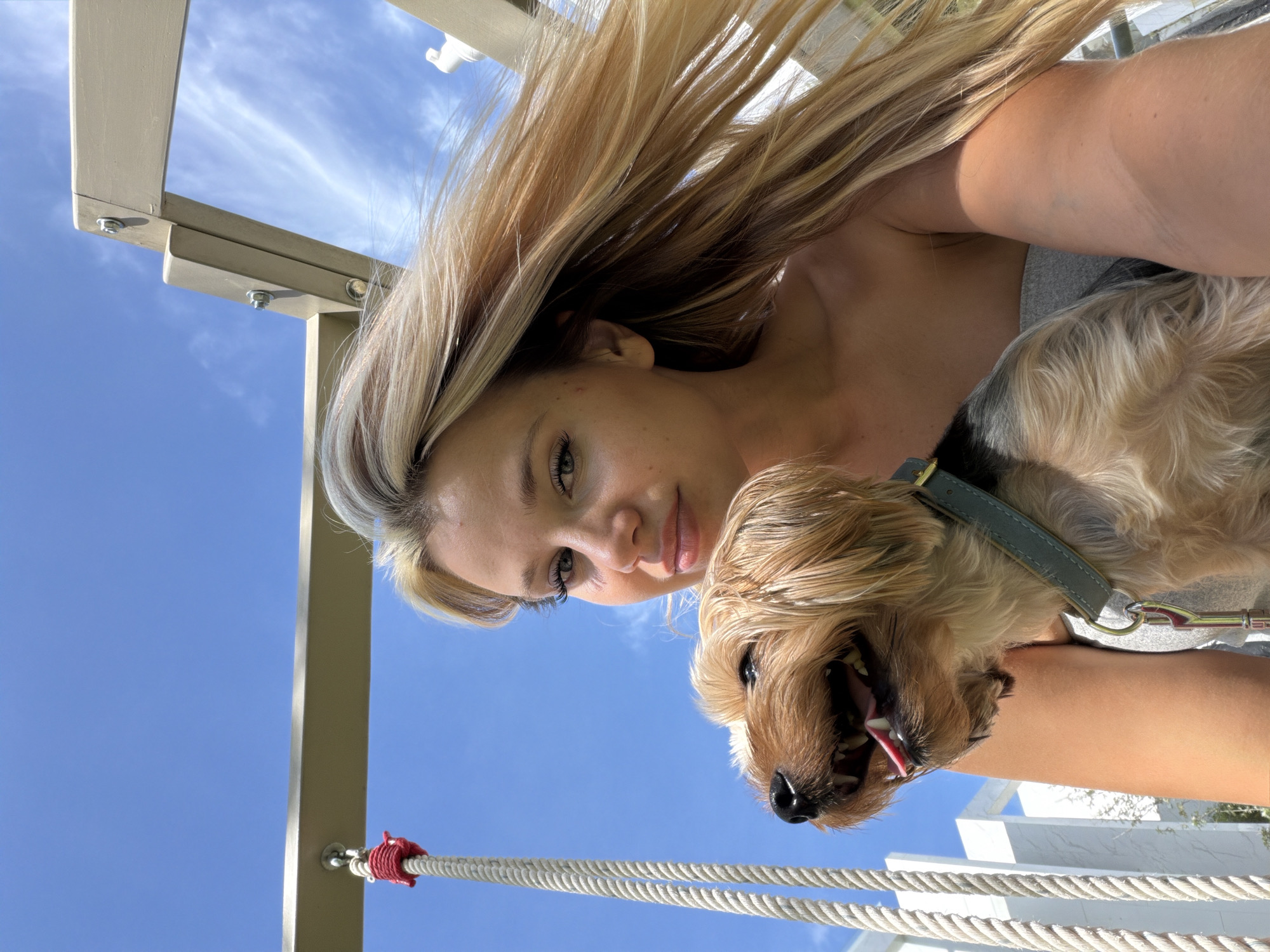 In this self-taken photograph, a young woman with long, blonde hair is pictured outdoors with her small, brown dog. The woman is holding the dog, and both are looking towards the camera. The dog appears happy, with its mouth open and tongue visible. The woman's expression is gentle, and she is smiling slightly.
The background consists of a bright blue sky with scattered, wispy clouds. Parts of a light-colored structure, possibly a swing set or playground equipment, are visible in the upper left and lower right of the frame. A thick rope, tied with red cord, runs across the lower portion of the image.
The lighting suggests it is daytime, likely sunny, with the sun casting a warm glow on the woman's arm and face. The overall mood is casual and joyful, capturing a moment of connection between the woman and her pet in an outdoor setting. The location context provided is Thep Krasatti, Thailand, though no specific visual cues within the image directly indicate this location. There is no visible text in the image.