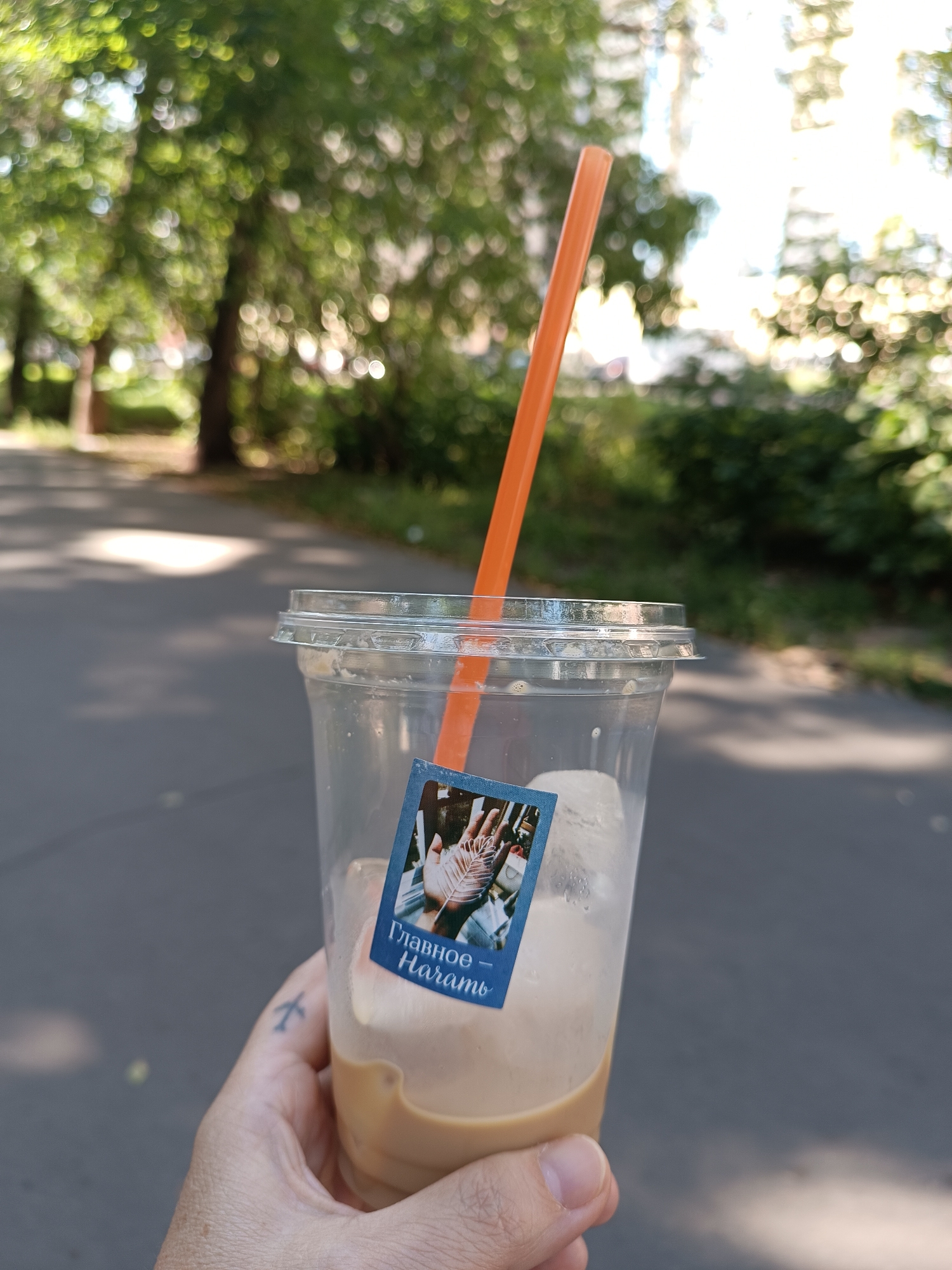 An outdoor medium shot captures a person's hand holding a clear plastic cup containing an iced coffee drink, with a bright orange straw protruding from the lid. A small blue sticker is affixed to the cup, featuring an image and text that reads "Главное – Hачать" (which translates to "The main thing is to start"). The background shows a blurred view of trees lining a paved path, suggesting a park or street in the city of Zhukovsky, Russia, with sunlight filtering through the leaves. The person's hand has a small tattoo of an airplane on their index finger.