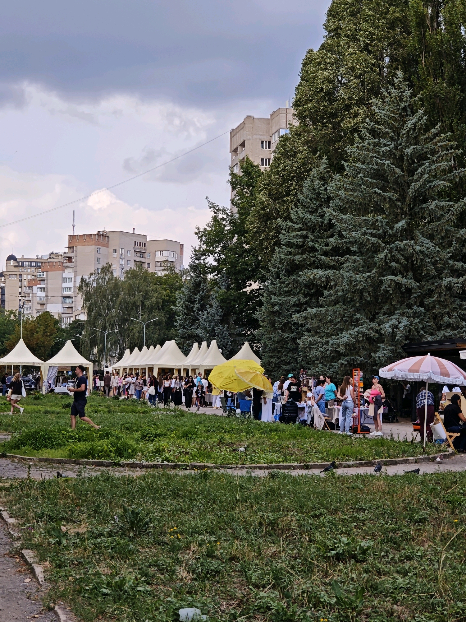 This image depicts an outdoor market or fair in Vinnytsia, Ukraine. In the foreground, there is a grassy area with some scattered debris and a paved pathway. Beyond this, a row of white tents, likely stalls for vendors, stretches across the scene. Several people are visible browsing or interacting at these stalls. Among the tents, a bright yellow umbrella stands out. In the background, a line of mature evergreen trees forms a backdrop, with several multi-story apartment buildings visible behind them. The sky is overcast with clouds. A man in a black t-shirt and dark shorts is walking in the middle ground.