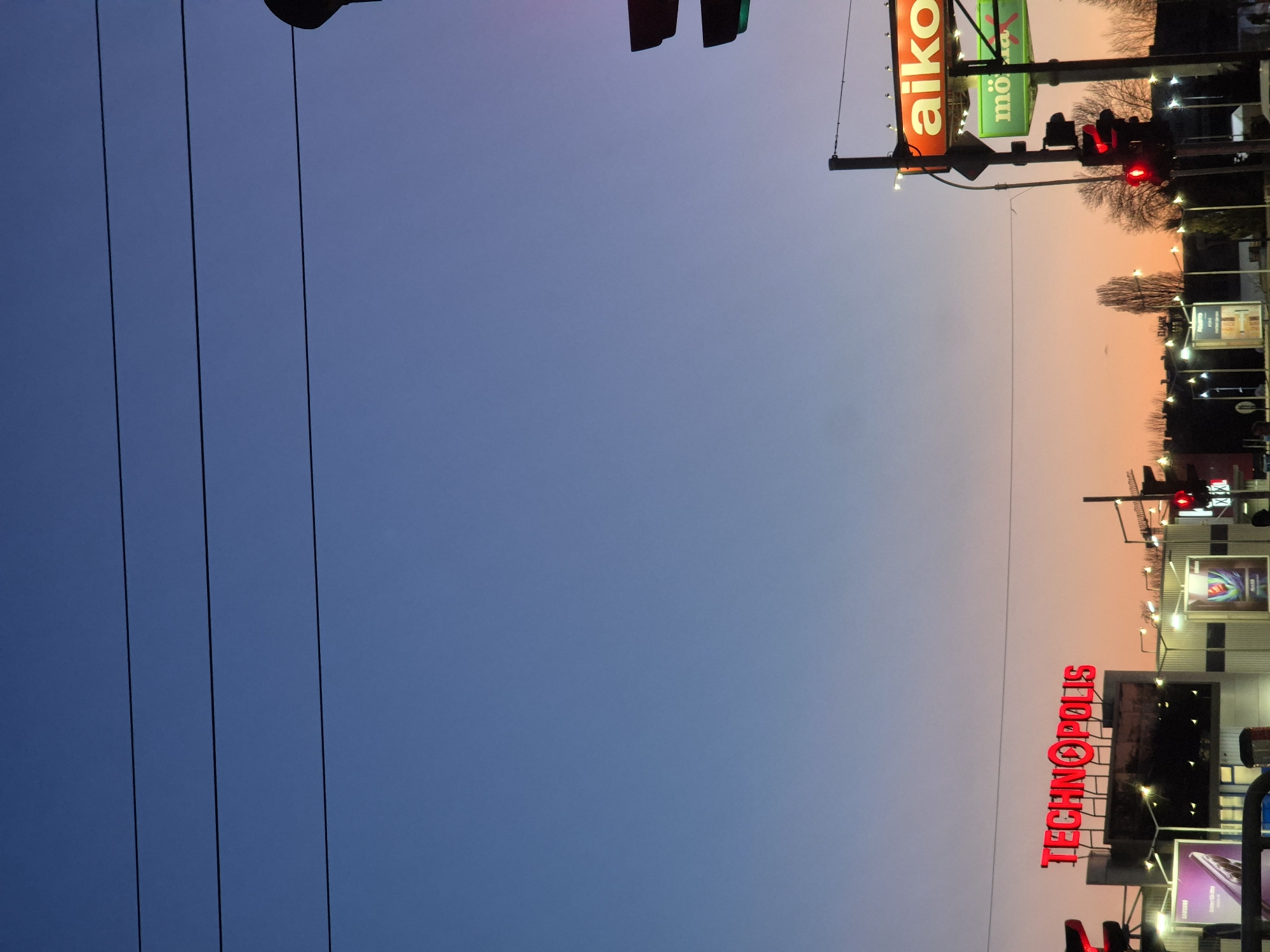 The image depicts a street scene in Varna, Bulgaria at dusk. The sky transitions from blue to orange. Visible on the right side is the "TECHNOPOLIS" store with a bright red sign. Further up the street are retail stores "Aiko" and "Mömax" with signs illuminated. Traffic lights are visible, showing red. Power lines run across the left side of the frame.