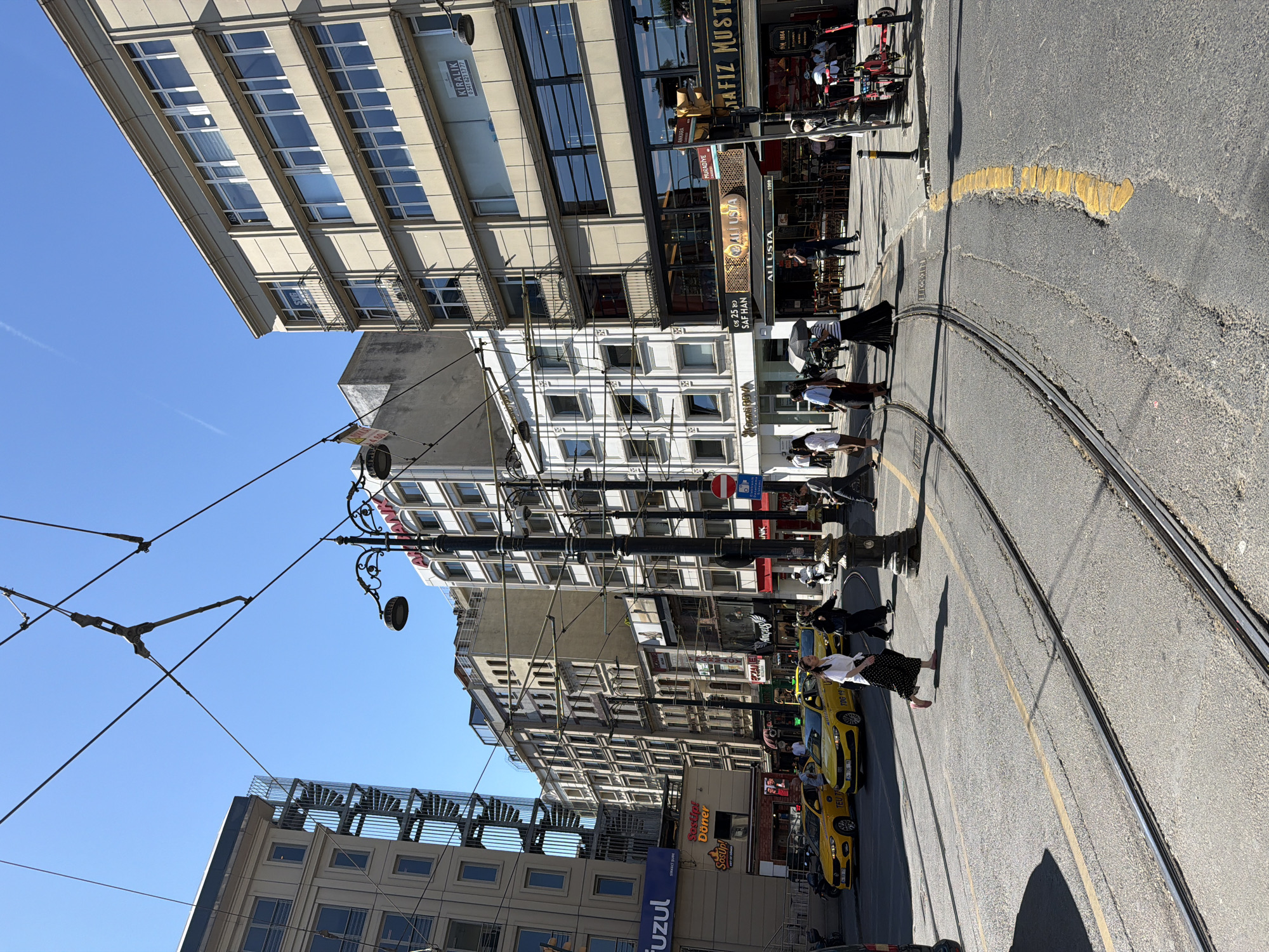 This is a photograph taken on a sunny day in Istanbul, Turkey, likely in the Beyoğlu district, given the presence of tram tracks and the architectural style of the buildings. The scene depicts a street intersection with tram lines running through it. Several yellow taxis are visible, indicating a busy urban environment. Pedestrians are crossing the street, with some walking and others holding umbrellas. The buildings lining the street are multi-story structures with a mix of modern and older architectural features. Overhead, power lines for the tram system are strung across the sky. The overall impression is of a vibrant and active city street.
