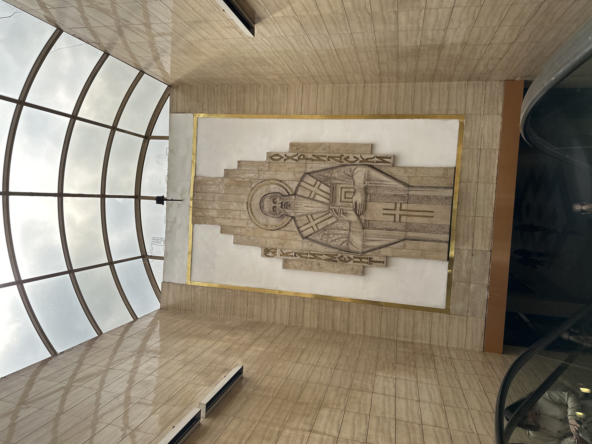 The original image, oriented 90 degrees counter-clockwise, depicts a modern-looking interior space dominated by a large, light-colored stone relief sculpture. The sculpture portrays a religious figure, likely an Orthodox saint, with a halo, beard, and ecclesiastical robes featuring cross motifs. The saint is shown holding a book or scroll. Cyrillic text carved into the stone surrounding the figure identifies it as "СВЕТИ КЛИМЕНТ ОХРИДСКИ" (Saint Clement of Ohrid), with "ОХРИДСКИ" visible above the head and "КЛИМЕНТ" to the side. The relief is framed by a thin gold border and set against a background of light-colored stone and white plaster.
To the left, a large, arched glass ceiling or window allows abundant, diffused natural light to enter, revealing an overcast sky outside. The interior walls and floor are covered in light-colored, vertically oriented rectangular tiles, creating a consistent and textured architectural aesthetic. On the far right, a dark, reflective, and curved surface, possibly part of an escalator or a large architectural element, is partially visible. The scene is static, showcasing the blend of modern architecture and religious art within what appears to be a public or institutional building, such as a church, museum, or university building in Sofia, Bulgaria. The overall atmosphere conveys solemnity and grandeur.