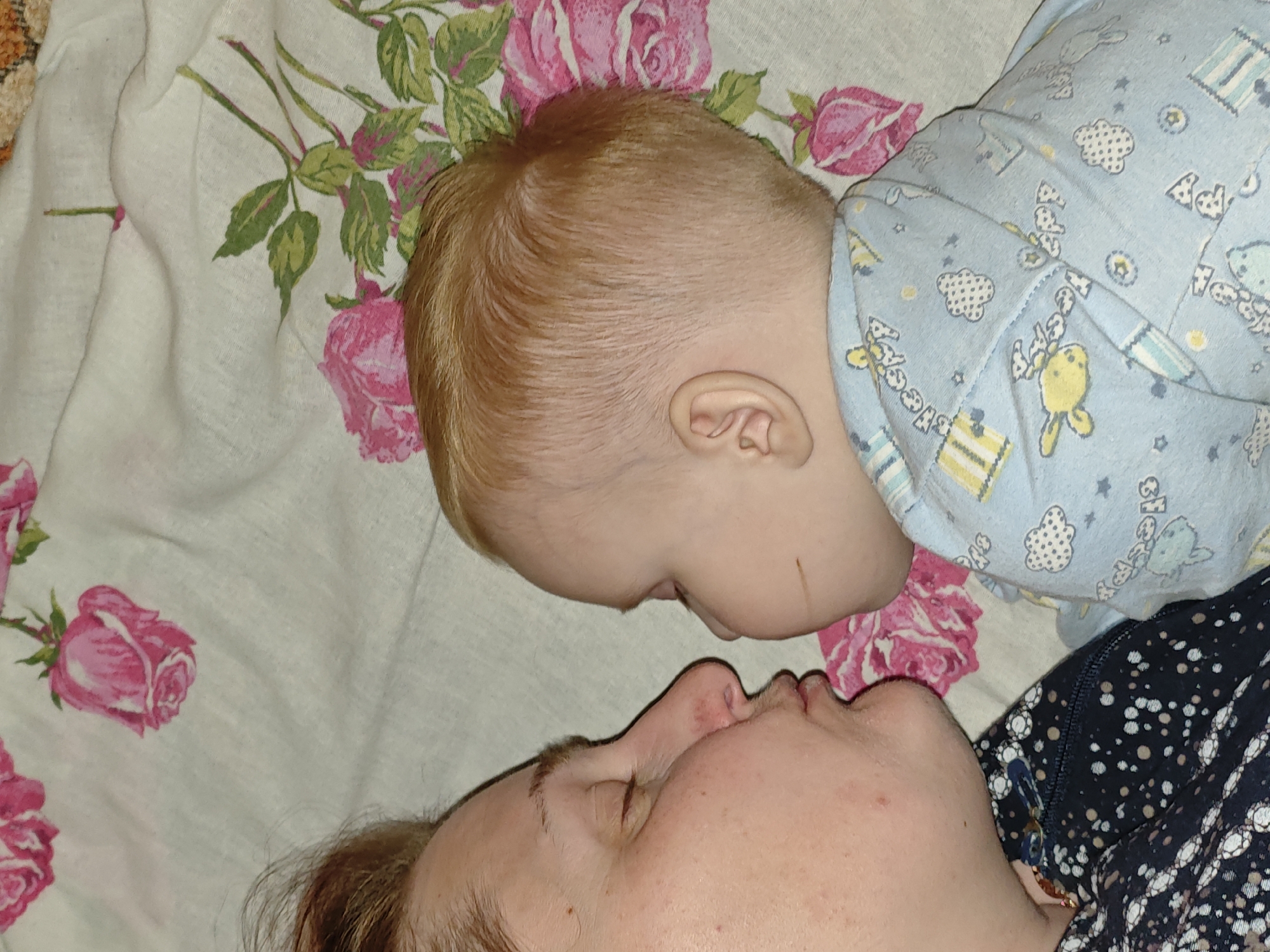 A woman and a baby are lying on a floral-patterned surface. The baby, wearing a blue patterned onesie, is positioned above the woman, with their faces close together. The woman has her eyes closed, while the baby's face is partially visible. The floral pattern on the surface consists of pink roses and green leaves. The location is Фоки, Russia.