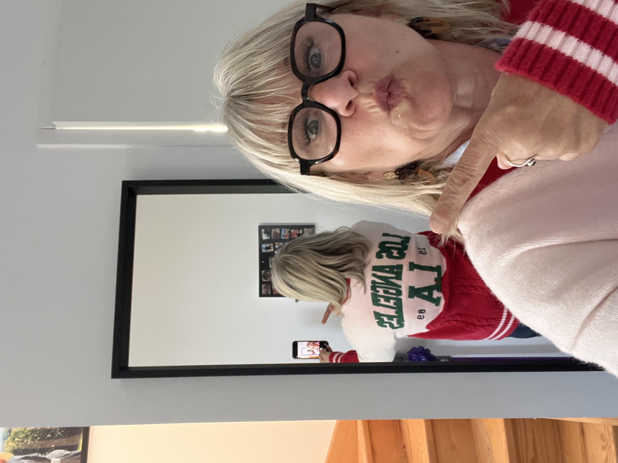 A woman with blonde hair and glasses is taking a selfie in a mirror. She is wearing a red and white striped sweater over a white shirt, and her finger is pointing towards her shoulder. The reflection in the mirror shows her back, wearing a white sweater with "LA" in green lettering. Behind the mirror, there is a wall with a photo collage. The scene appears to be indoors, with a hint of wooden stairs visible in the lower part of the image. There is no clear indication in the image that locates it in Chambœuf, France.