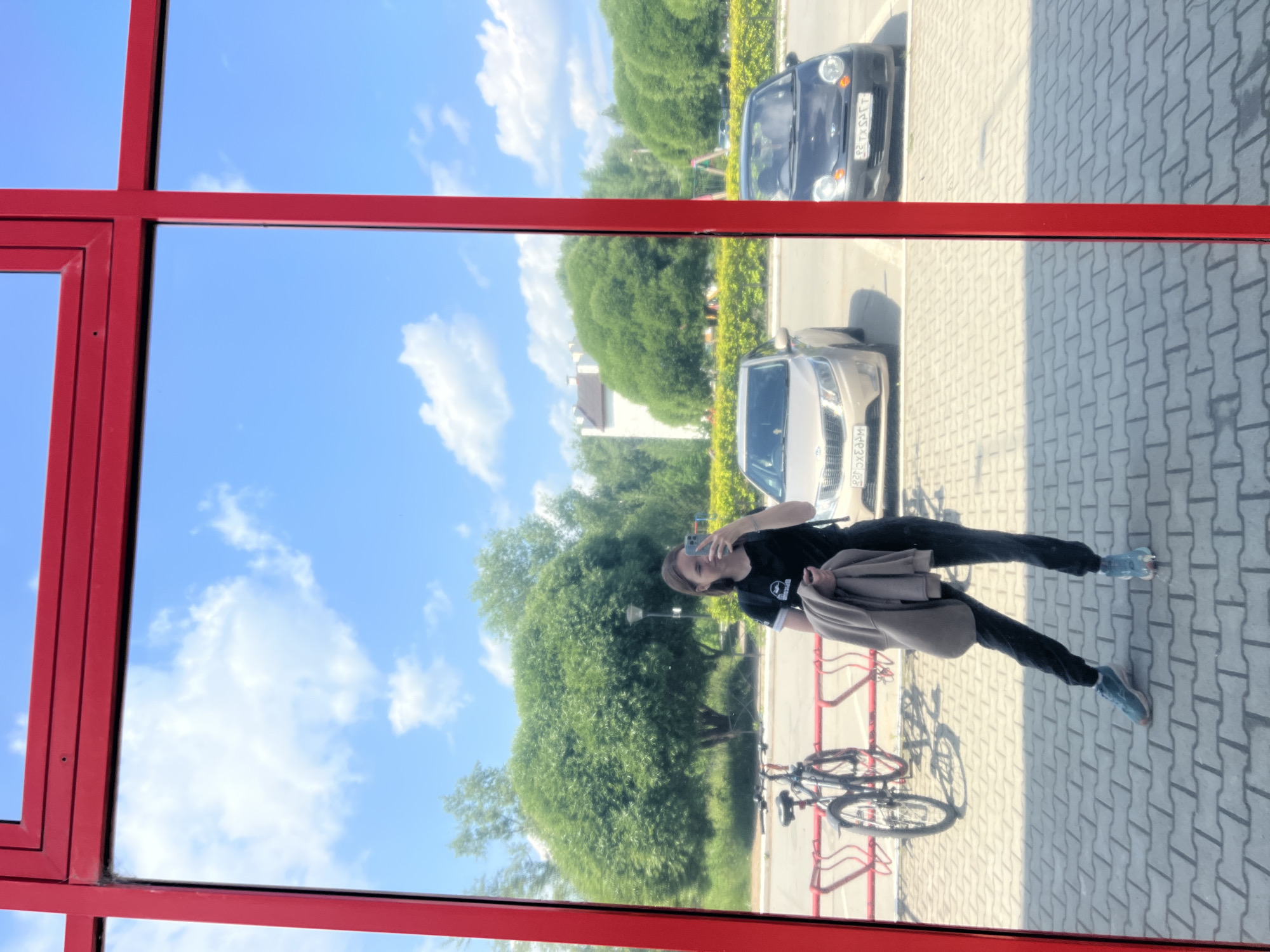 A young woman is taking a mirror selfie in front of a building with a bright red frame. She is wearing a black t-shirt, dark pants, and light blue sneakers, and holding a light brown jacket over her arm. In the reflection, a bicycle is parked at a bike rack to her left. Behind her, two cars are parked in a lot, with a row of green bushes and trees in the background. The sky is blue with scattered white clouds. The scene appears to be in a public outdoor space, possibly near a shopping area or residential building, in the city of Chaikovsky, Russia.