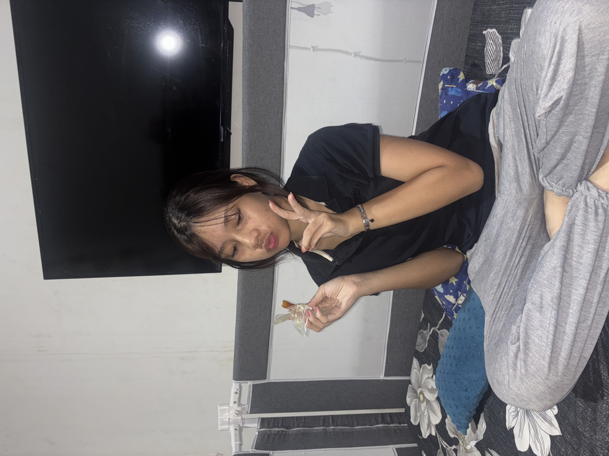 A young woman of Southeast Asian descent, with dark hair and brown eyes, is seated on a bed in a room. She is wearing a black polo shirt and grey sweatpants. In her right hand, she holds a small, wrapped item, possibly food or candy. Her left hand is raised, with two fingers extended in a "peace" gesture. She is looking towards the item in her hand with a playful expression. The bed is covered with a dark floral patterned sheet and there are several pillows on it. A large flat-screen television is mounted on the wall behind her. The wall behind the bed is off-white with visible water stains. The room appears to be a bedroom. The overall scene suggests a casual, indoor setting.