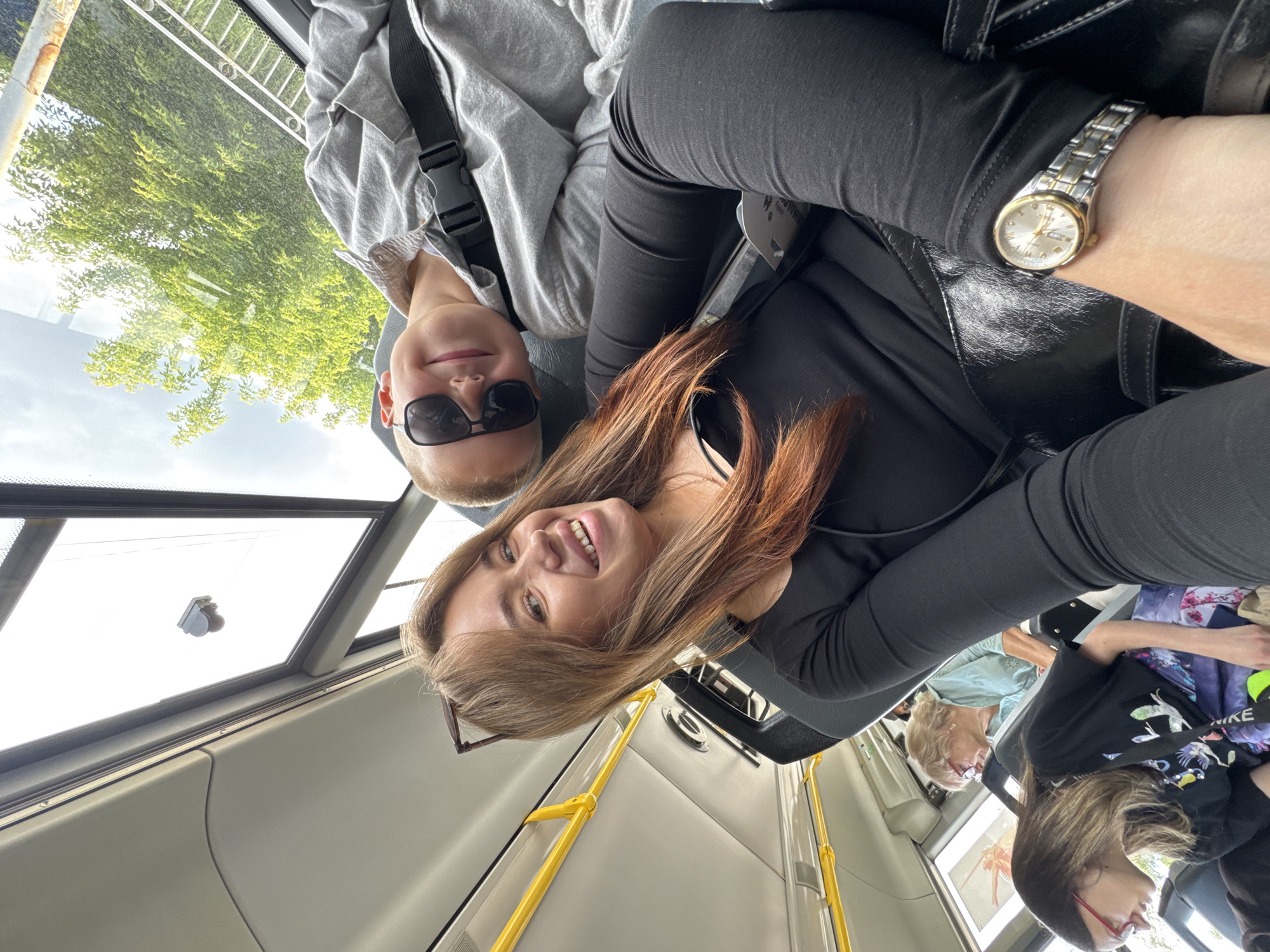 In this selfie-style photograph taken from inside a vehicle, likely a bus or train, two women and a young person are visible. The primary subjects in the foreground are a woman with long, reddish-brown hair and a young person wearing sunglasses and a grey hooded jacket. The woman is smiling directly at the camera, and the young person is also looking towards the lens. To the right of the woman, a wrist with a silver and gold watch is in view.
In the background, two other women are seated. One, with blonde hair and glasses, is wearing a light blue top. The other, with brown hair and glasses, is wearing a black t-shirt with a colorful graphic print. The scene appears to be set during the daytime, with natural light filtering in from the windows. The windows show greenery and trees outside, suggesting an outdoor setting. The interior of the vehicle has cream-colored walls and yellow grab rails. No explicit text is visible in the image, though a tag can be seen on the young person's clothing. The overall mood of the photograph appears casual and friendly, with the smiling subjects in the foreground. The location context is Chaikovsky, Russia.