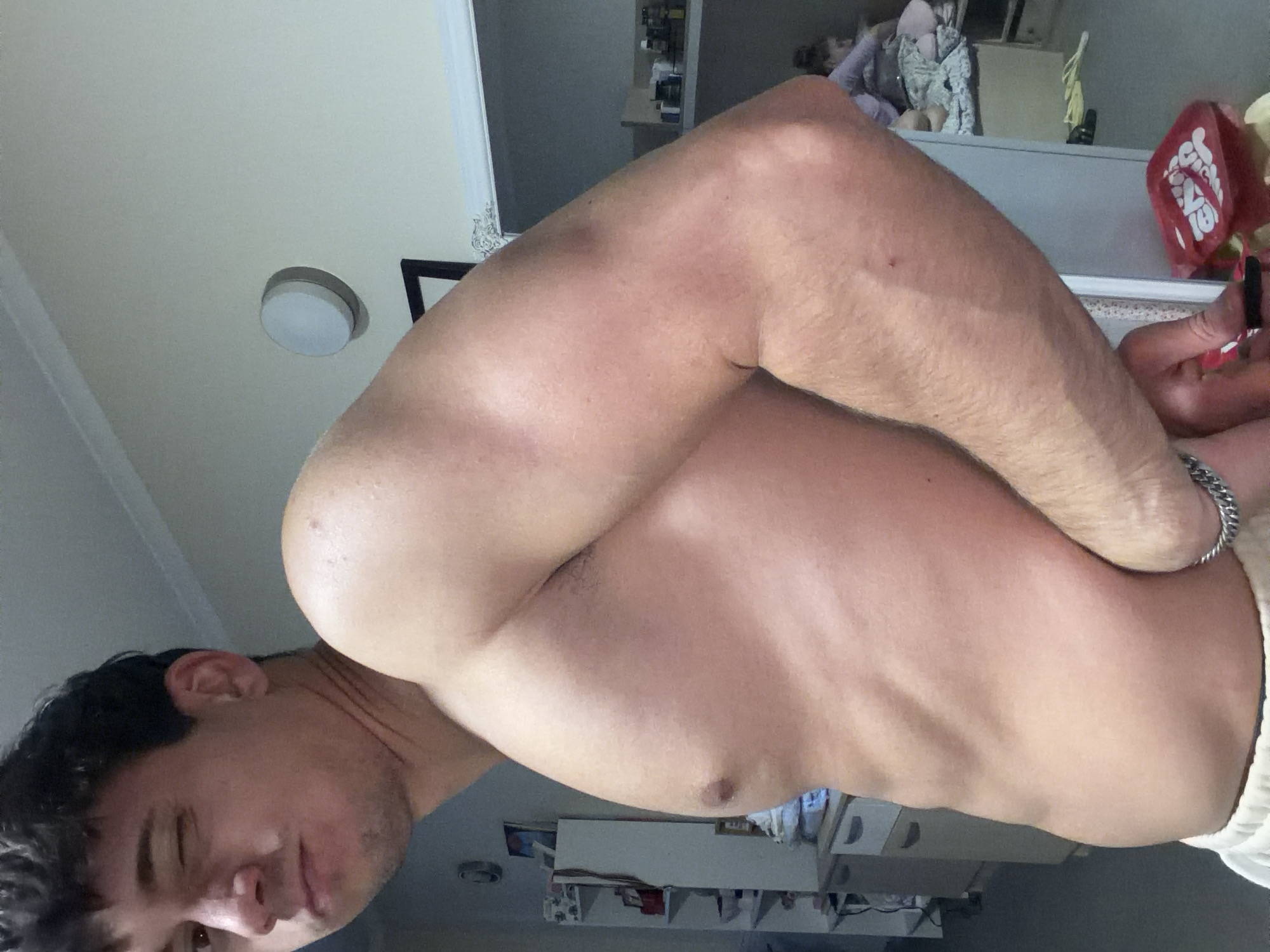 The original image is rotated 90 degrees clockwise, showing a young man indoors, likely in Sofia, Bulgaria. The man, who appears shirtless, is positioned in the foreground, filling much of the frame from the chest up. He has dark, wavy hair and is looking towards the camera with a neutral, slightly serious expression. His left arm is prominently featured, muscular and bent, with a silver chain bracelet visible on his wrist. His right arm is bent and appears to be behind his back or holding something out of frame. The man's torso is visible, showing a lean, muscular build with some chest hair and visible veins on his arm. White shorts or sleepwear are visible at the very bottom right of the frame.
The background is an indoor setting with various household items. In the upper left, a white ceiling with a round, recessed light fixture is visible. Below that, a reflective surface, possibly a mirror or cabinet door, is present. In the mid-ground and background, there are shelves or cabinets with various items on them. On the right side of the image, resting on a surface, is a red object that appears to be a dice cup or game accessory, with white text partially visible, including phrases like "GIVE IT A GO" and "GAME ON" (inverted due to the image orientation). Further in the blurred background, another person, possibly a child, can be seen lying down or resting on what looks like a bed or couch, partially covered by patterned fabric. The overall lighting suggests an indoor setting, possibly with artificial light sources.