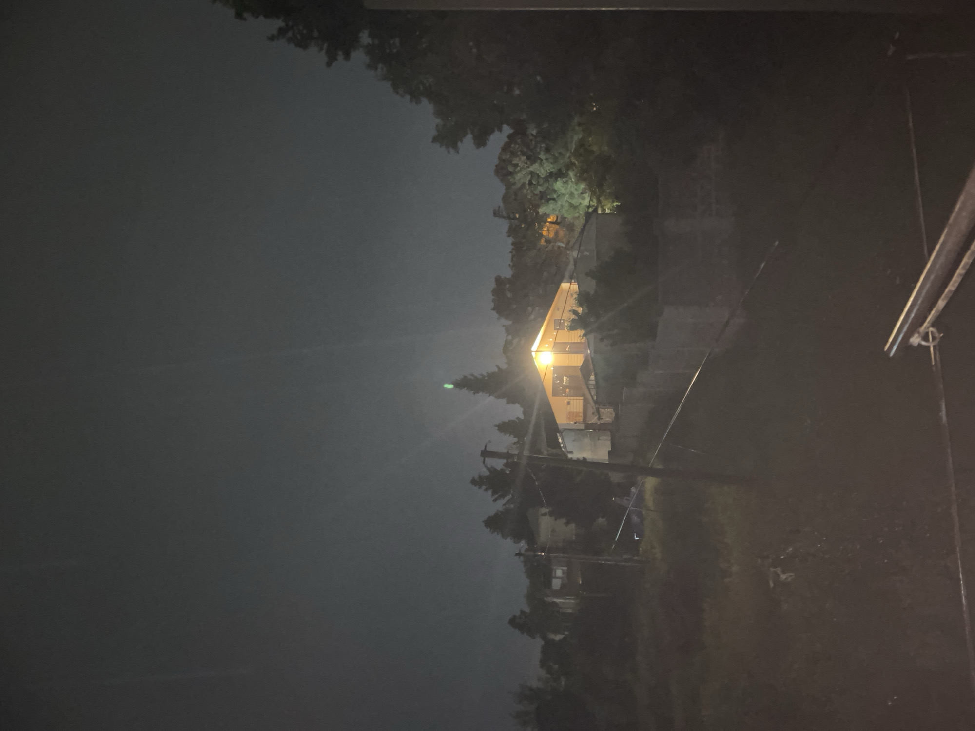 An exterior nighttime shot captures a dimly lit scene in Tbilisi, Georgia. A house, illuminated by warm interior lights that shine through its windows, is visible in the mid-ground. The house is surrounded by trees and other structures, some of which are partially obscured by darkness. A few utility poles with wires are also present. The sky above is dark with some atmospheric haze.