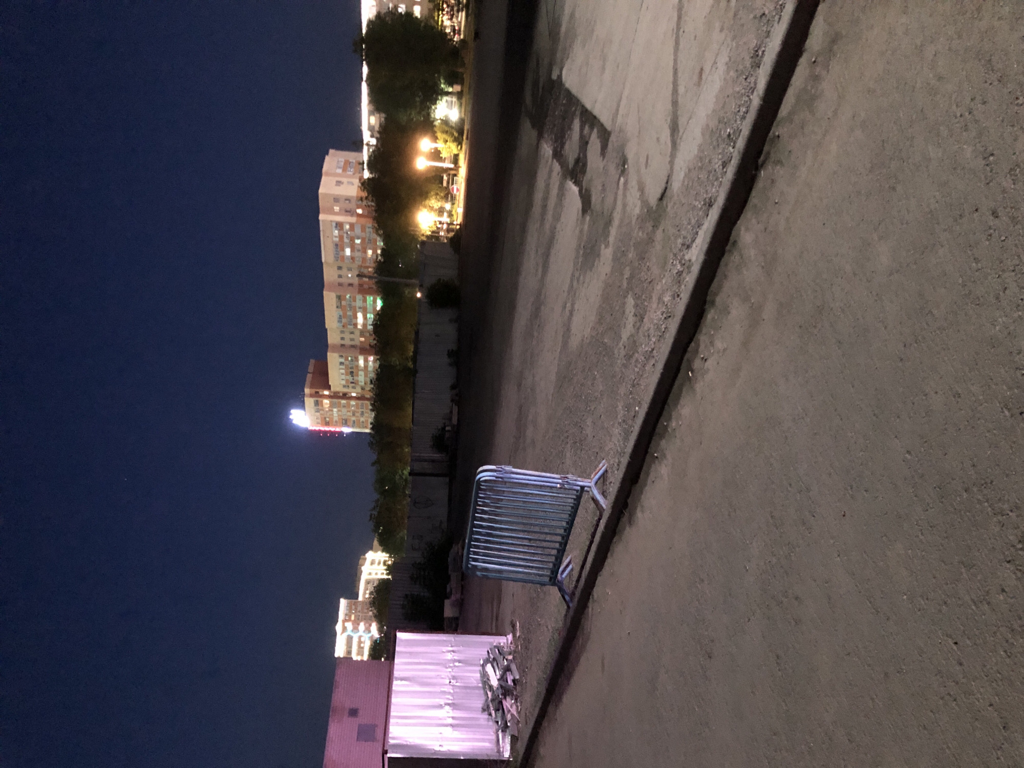 The image depicts an outdoor scene at dusk or night, with a dark blue sky. In the foreground, a paved area transitions to a gravelly surface near a curb. A blue metal barricade stands on the gravel, angled towards the left. To the left of the barricade, a light-colored, corrugated metal structure, possibly a shed or container, is visible, with a pile of metal debris in front of it. In the background, several multi-story buildings with illuminated windows are silhouetted against the sky, suggesting an urban environment. Trees line the area in front of the buildings. Streetlights are visible, casting warm glows. The overall impression is of an outdoor space in an urban setting after dark.
