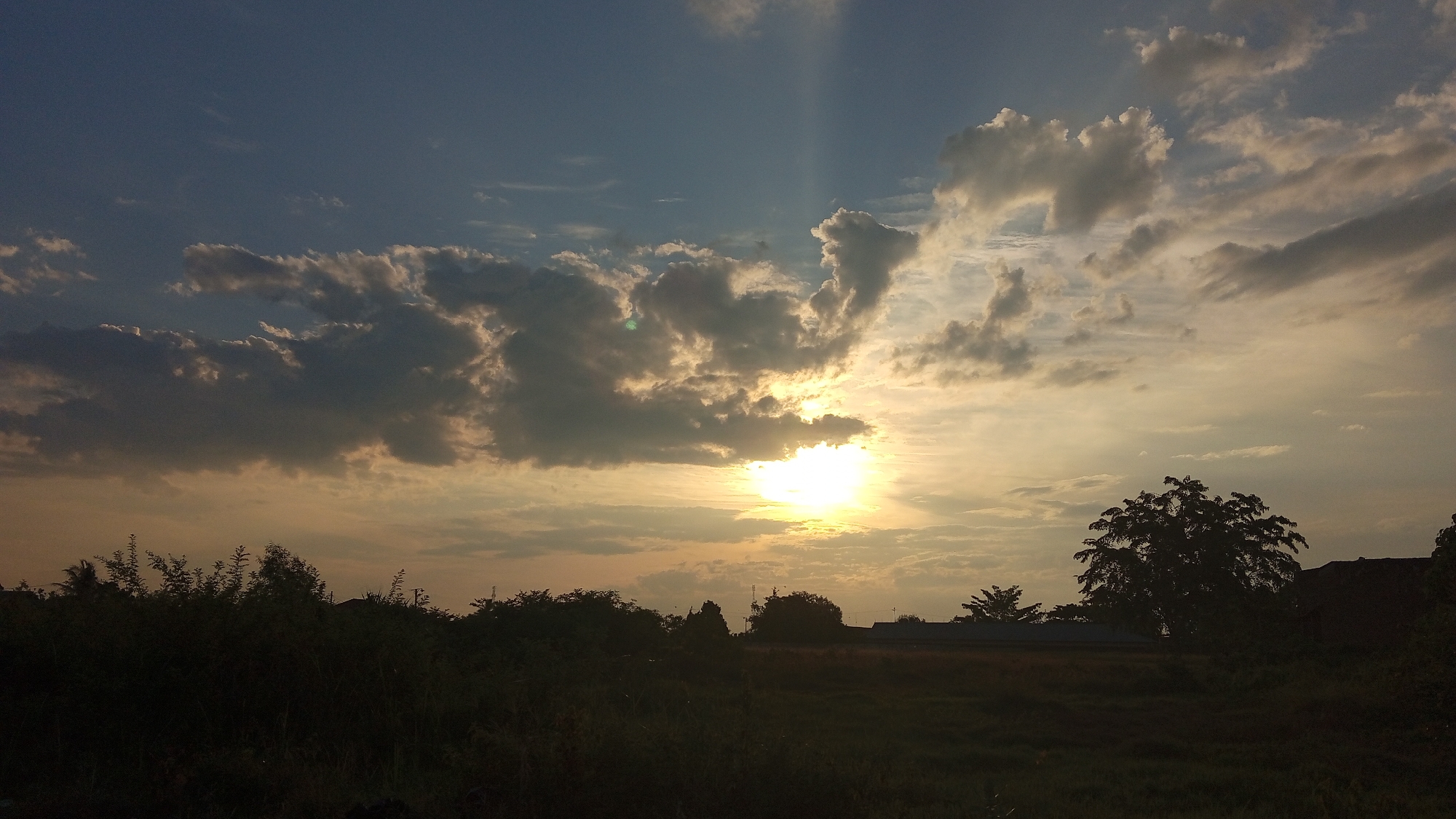 The image depicts a sunset scene in Uteunreutoh, Indonesia. The sky is filled with a mix of dark and light clouds, with rays of sunlight breaking through, illuminating the sky with warm hues of yellow and orange. The sun is visible as a bright, intense orb near the center of the frame, partially obscured by clouds. Silhouetted against the sky are various trees and the outlines of buildings in the distance. The foreground is dominated by dark, dense foliage and grassy areas, also rendered in silhouette due to the low light conditions. The overall impression is one of a natural landscape bathed in the soft, dramatic light of sunset.