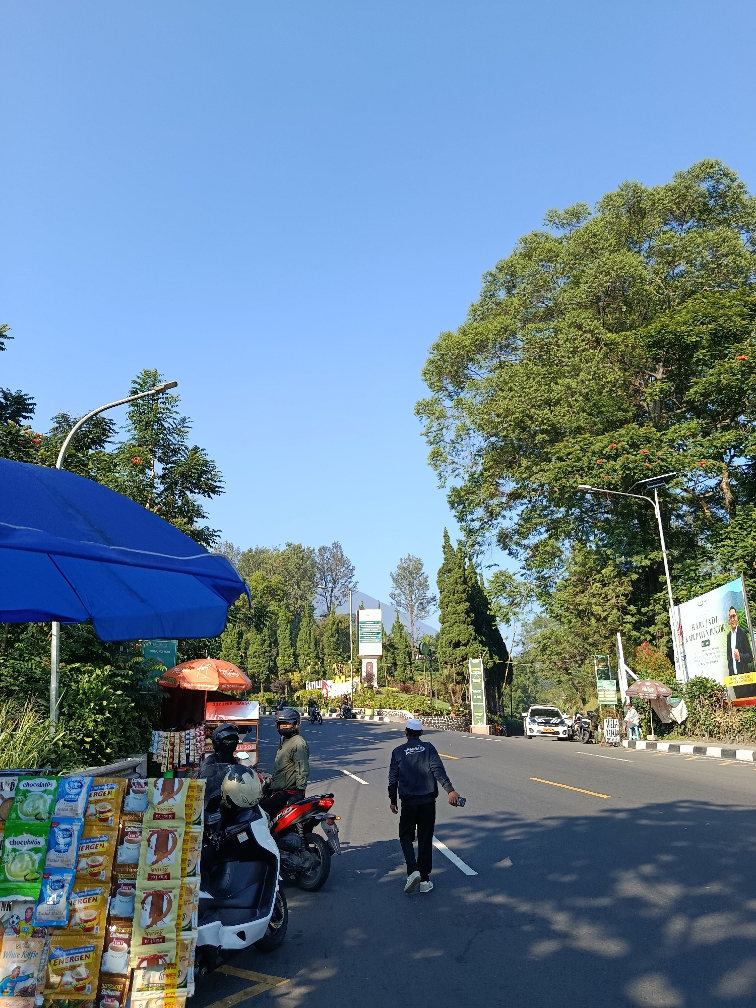 This is a street-level, medium-distance, eye-level, full-shot photograph taken on a sunny day in Cisarua, Indonesia. In the foreground, a stand is set up selling various packaged goods, including coffee and snacks, under a large blue umbrella. Several motorcycles are parked nearby, one of which has a person in a green jacket and helmet sitting on it, facing left. Another person, wearing a dark jacket with a white logo and a white cap, walks away from the camera on the right side of the road. A white car with police markings is visible further down the road, accompanied by another motorcycle. The road is asphalt with yellow lines. The background features lush green trees and some signs, including a billboard with a picture of a man. The sky is clear and blue. The scene depicts a commercial roadside area in a town with a clear, bright atmosphere.
