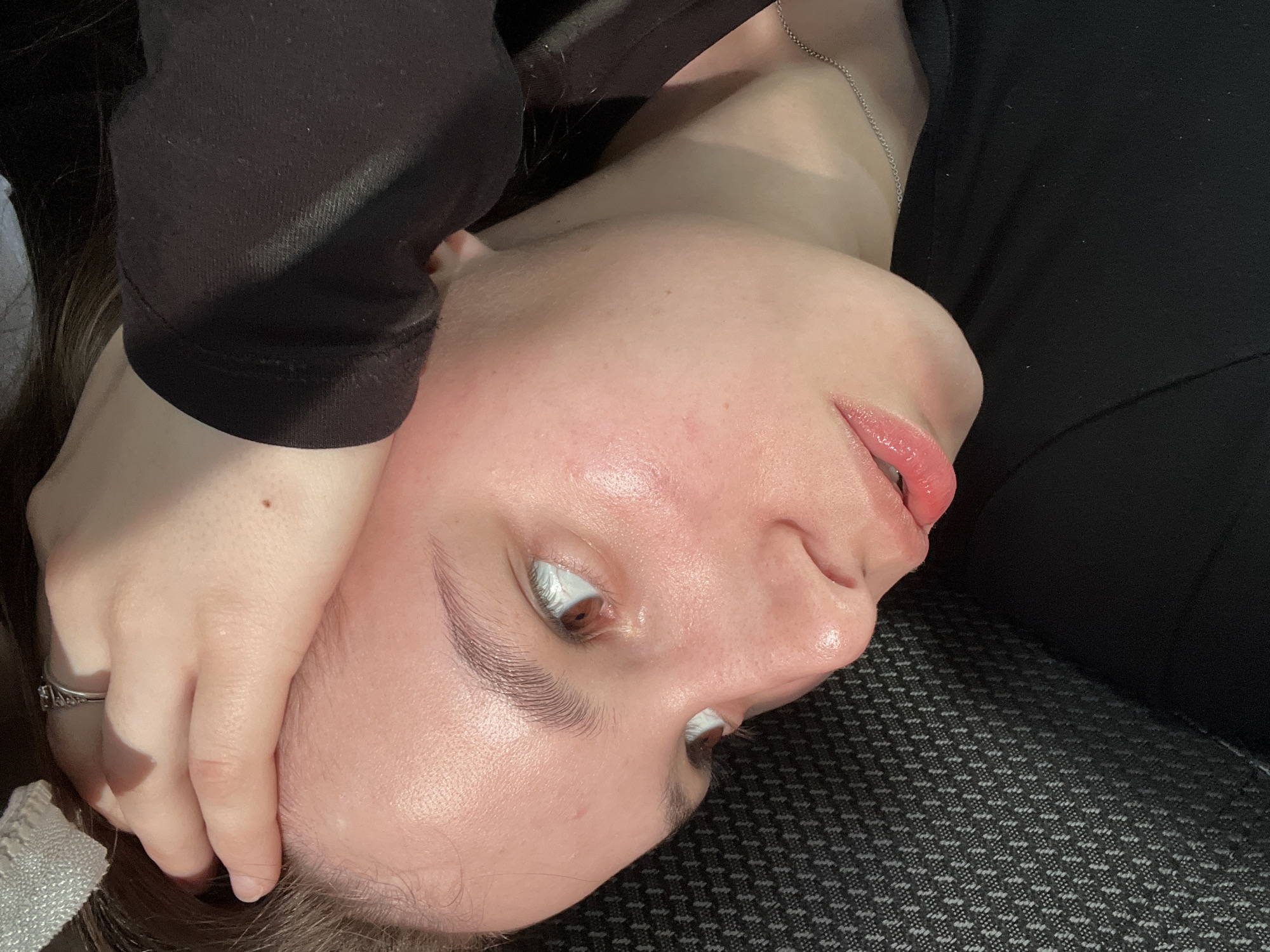 A close-up, angled shot of a person's face with light skin. The person's head is resting on what appears to be a dark grey textured surface. The person's right hand is positioned on their forehead. The person is wearing a black top. A silver necklace is visible around the neck. The location is unknown.