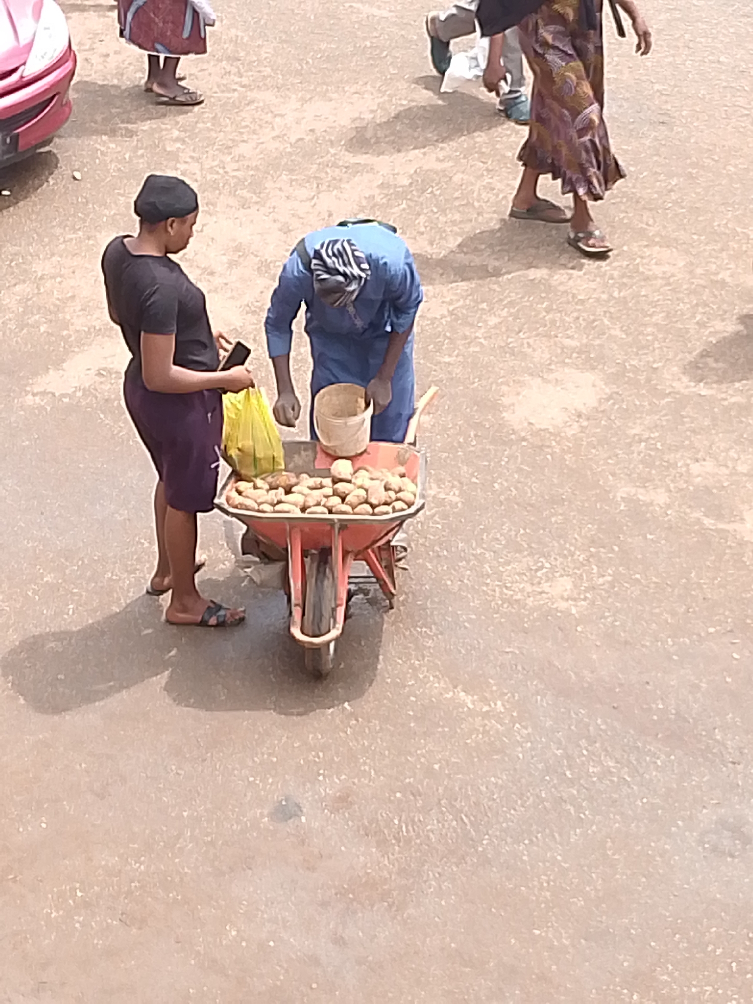 In Bukuru, Nigeria, a street scene unfolds with two women and a wheelbarrow laden with produce. One woman, wearing a black t-shirt and purple shorts, stands to the left, holding a yellow plastic bag in her right hand and a smartphone in her left. Her feet are clad in black and brown sandals. To her right, another woman in a blue kaftan and headscarf leans over the wheelbarrow, which is filled with potatoes. She is holding a small brown bucket. The wheelbarrow itself is red and has a single wheel. The ground is a sandy, outdoor surface. In the background, a portion of a pink car is visible on the far left, and another person in a patterned skirt and sandals stands further back. The overall impression is a bustling marketplace environment.