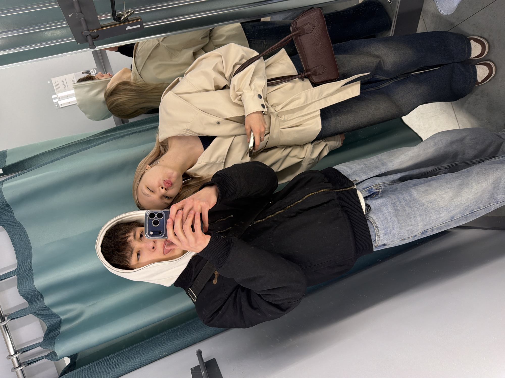 The image, taken in Yekaterinburg, Russia, depicts two young individuals posing for a mirror selfie in what appears to be a fitting room. The photo is displayed in a landscape orientation, but the subjects are standing upright, captured in their reflection.
In the foreground and closer to the camera, a person with short dark hair, wearing a white hooded sweatshirt underneath a black zippered jacket and light blue jeans, holds a dark-colored smartphone to capture the reflection. Their face is visible in the phone's screen, showing a playful expression with their tongue slightly extended.
Behind them, a second person with blonde hair is reflected in the mirror. They are dressed in a light beige trench coat, dark blue jeans, and brown and white sneakers. This individual holds a silver smartphone and displays a relaxed, slightly pouting expression. A dark brown shoulder bag is slung over their arm.
The background reveals a large mirror, a light blue-green textured curtain to the left, and a light gray floor. Above the curtain, a metal rail and a small black tag with a partially visible white number, likely "1," are reflected, indicating a fitting room number. An unreadable white sign is also visible in the reflection above the blonde person's head. The scene captures a casual moment of friends or companions trying on clothes or taking a fun photo.
