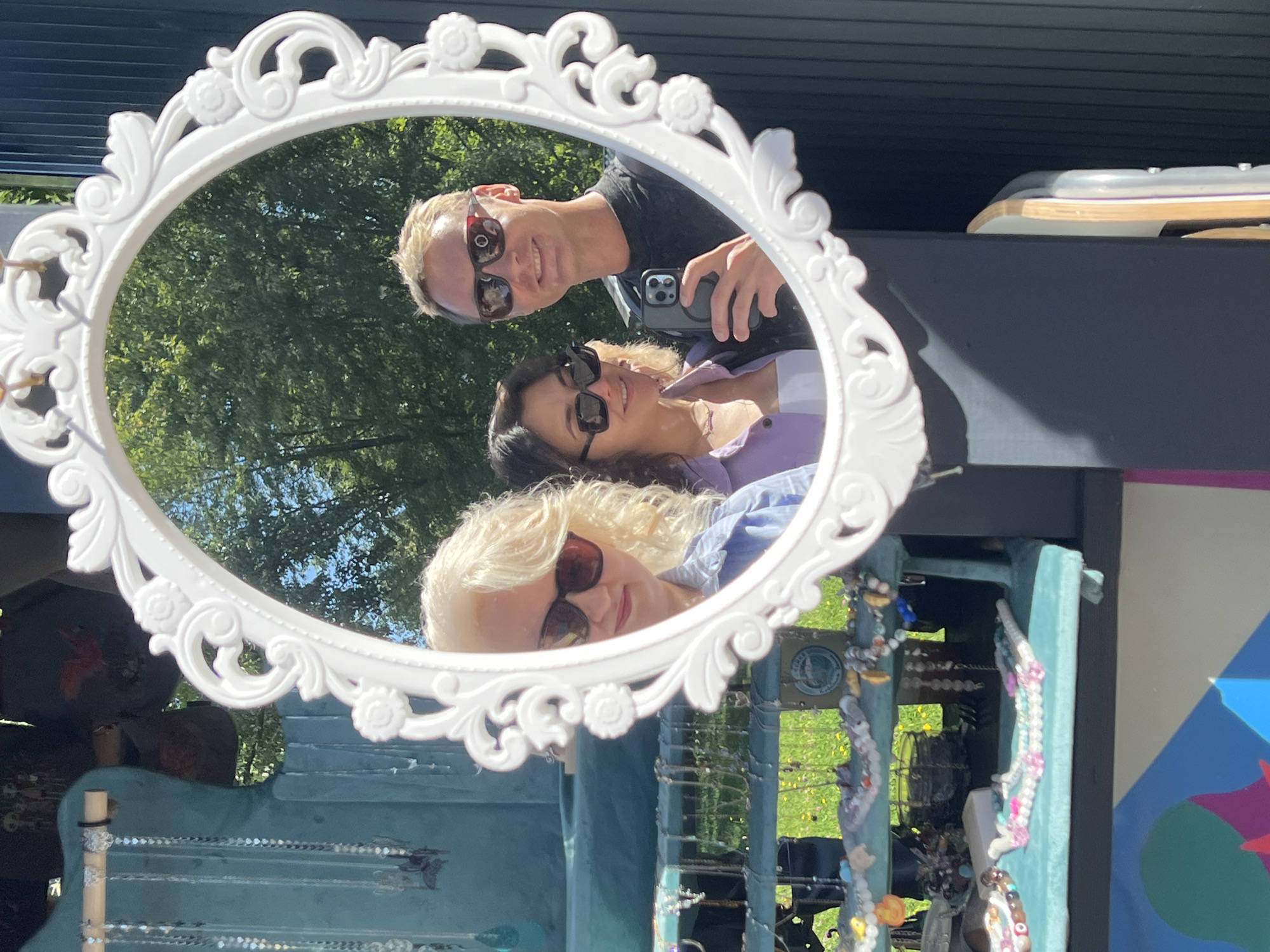 In Moscow, Russia, a mirror framed with ornate white detailing reflects three people. The person on the far left, a woman with blonde curly hair and sunglasses, is wearing a blue shirt. Next to her, a woman with dark hair and sunglasses is wearing a purple shirt over a white top. On the right, a man with blond hair and sunglasses is holding a smartphone, wearing a black t-shirt. They appear to be at an outdoor market, with jewelry displayed on teal-colored stands behind them. The background shows green trees and a blue sky, indicating an outdoor setting.