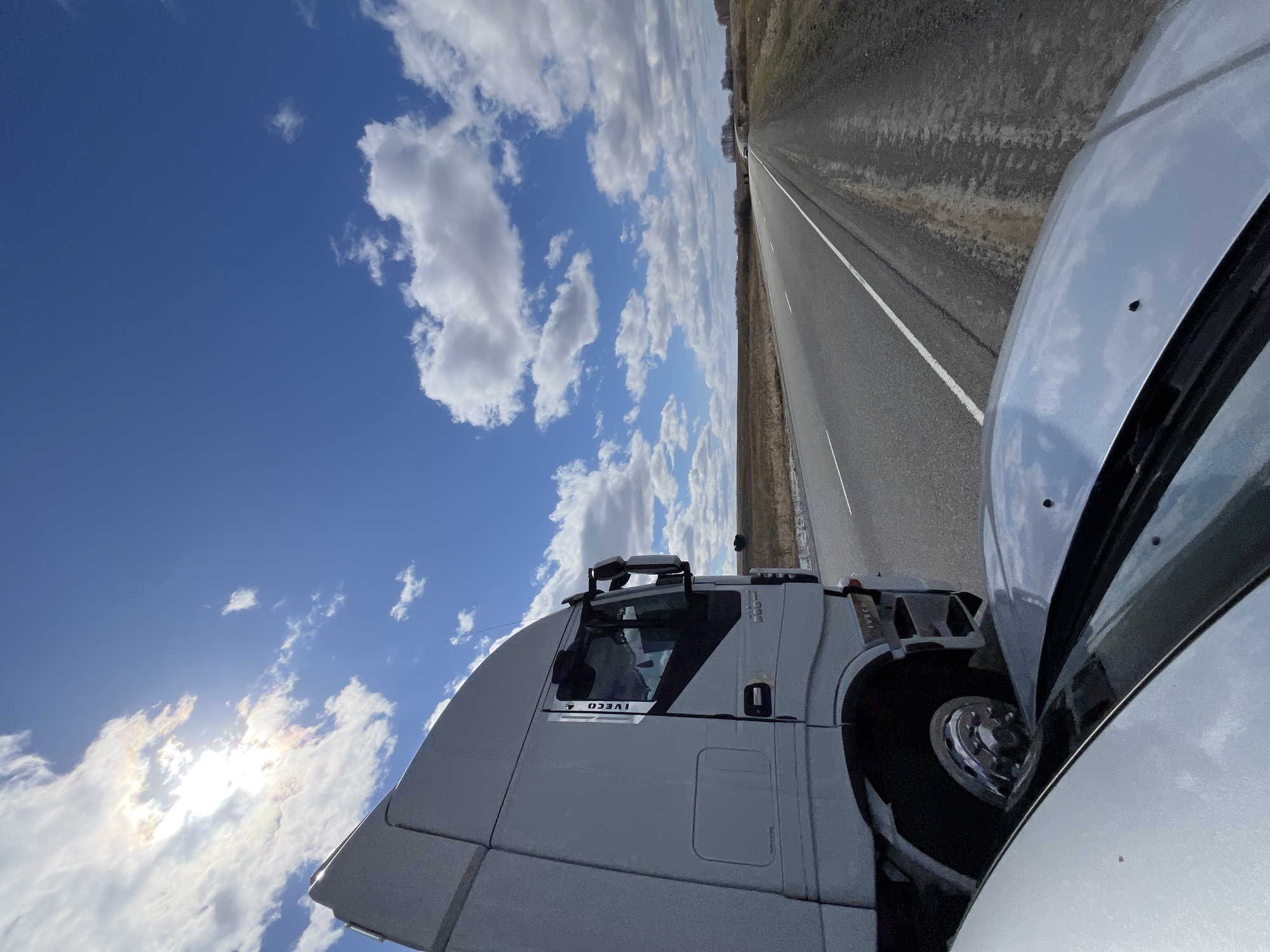 A low-angle shot shows a section of highway in Zhandat, Russia, under a bright blue sky with puffy white clouds. A large silver truck is parked on the side of the road. Another silver vehicle, potentially a car, is partially visible in the foreground. The landscape bordering the road is mostly dry vegetation. The highway stretches out in a straight line, converging in the distance.