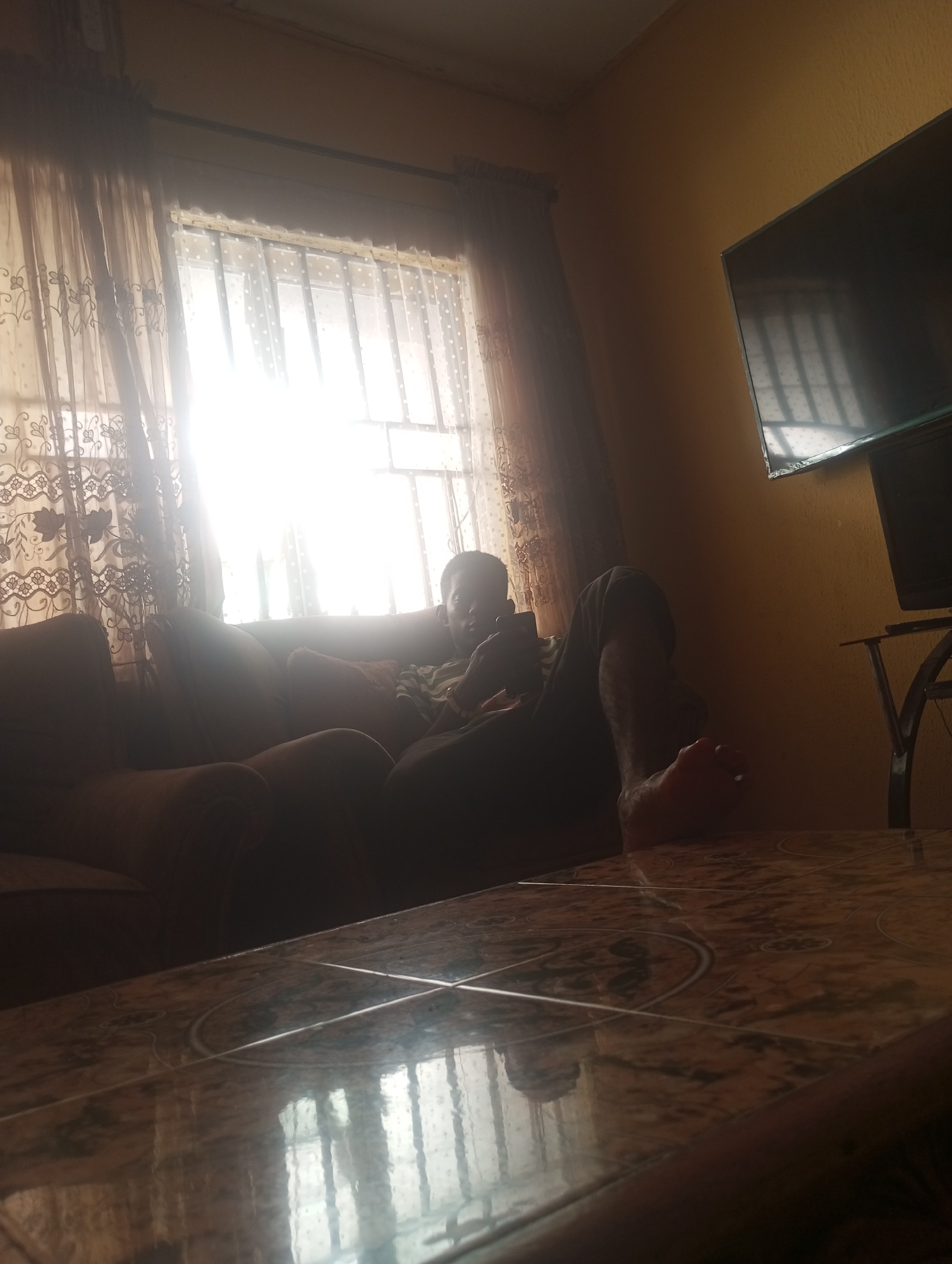 A low-angle, indoor shot captures a person relaxing on a sofa in a room. The person, positioned on the right side of the sofa, is partially visible, wearing a striped t-shirt and dark pants. Their legs are extended, with one foot resting on what appears to be a table. The room features a window with sheer, patterned curtains on the left, allowing bright sunlight to stream in, creating a strong backlight. A television is mounted on the wall to the right. The foreground is dominated by a tiled table, reflecting the light from the window and the room's interior. The overall scene suggests a casual, domestic environment, likely in the city of Onitsha, Nigeria, based on contextual information.