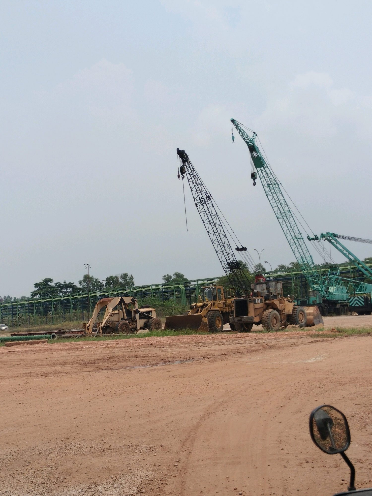 The image shows a construction site in Batam, Indonesia, with several pieces of heavy machinery, including cranes and front-end loaders. Two large cranes, one black and one green, dominate the background, with their booms extended. In the foreground, two yellow front-end loaders are parked side-by-side. To the left, a smaller, older piece of machinery, possibly a backhoe, is visible. In the far background, a long, green pipe structure runs horizontally, supported by metal scaffolding, and is surrounded by trees. The ground is composed of red dirt and gravel. The sky is overcast with light clouds. The image appears to be taken from the perspective of someone on a motorcycle, as a rearview mirror is visible in the lower right corner.