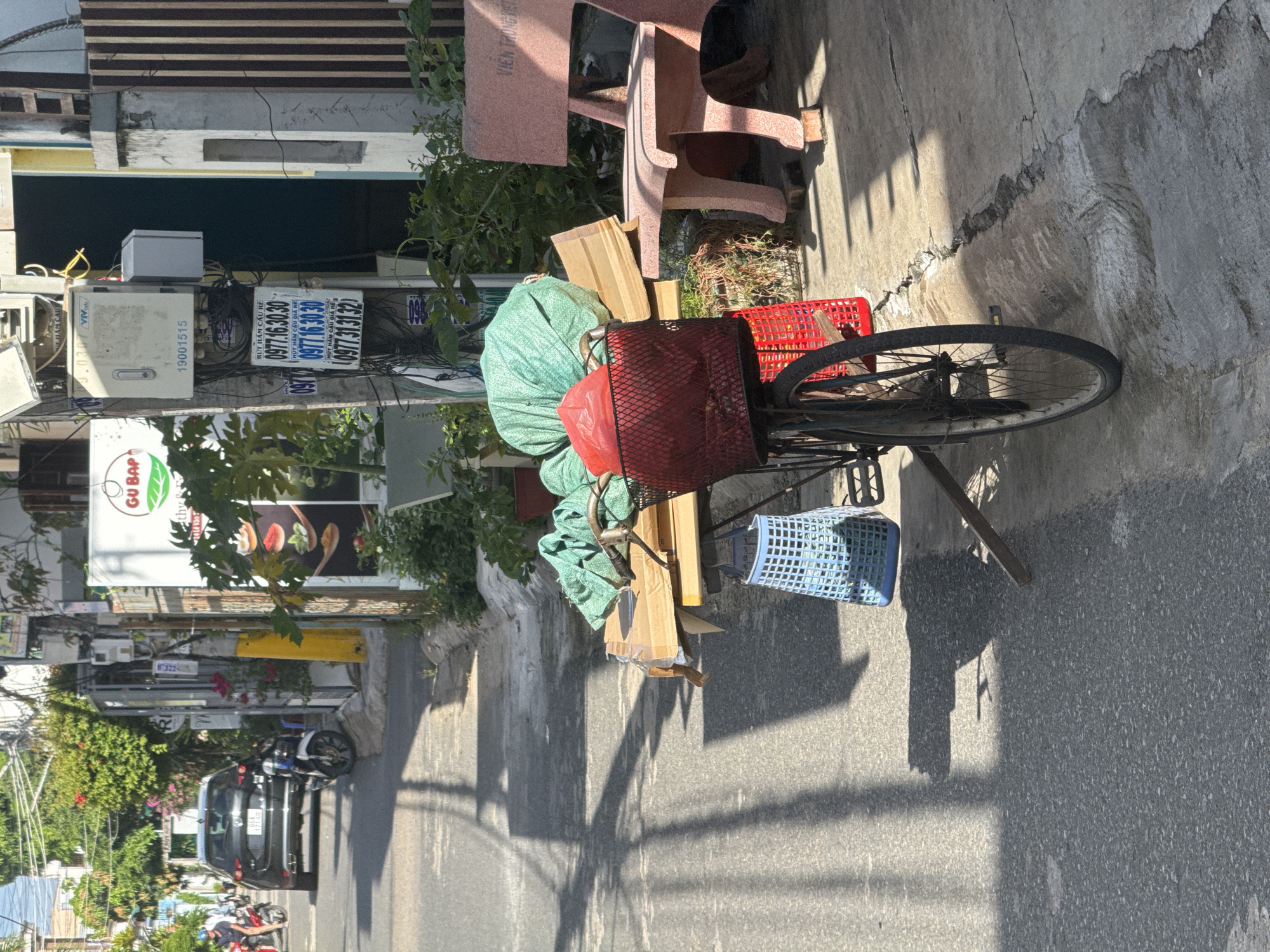 In the city of Phú Thủy, Vietnam, a street scene unfolds under bright sunlight, casting sharp shadows. The focus is on a bicycle loaded with various items. A large green tarp is bundled on the rear rack, topped with a red plastic bag. Beneath this, cardboard boxes are stacked precariously. A red wire basket holds more contents, and a blue plastic basket hangs from the bicycle's frame. The bicycle itself appears old, with a worn tire and visible spokes. To the right, a pink concrete bench with raised lettering that reads "VIỆN TRUNG TH" sits on the sidewalk. Behind the bench, the textured wall of a building is visible. The street pavement is cracked and uneven in places. In the background, a black car is parked on the left side of the street, and other motorcycles are visible. Buildings with awnings and greenery line the street, suggesting a commercial or residential area. Electrical wires and utility boxes are mounted on poles along the sidewalk. A sign with the text "GU BAP" is affixed to one of the buildings.