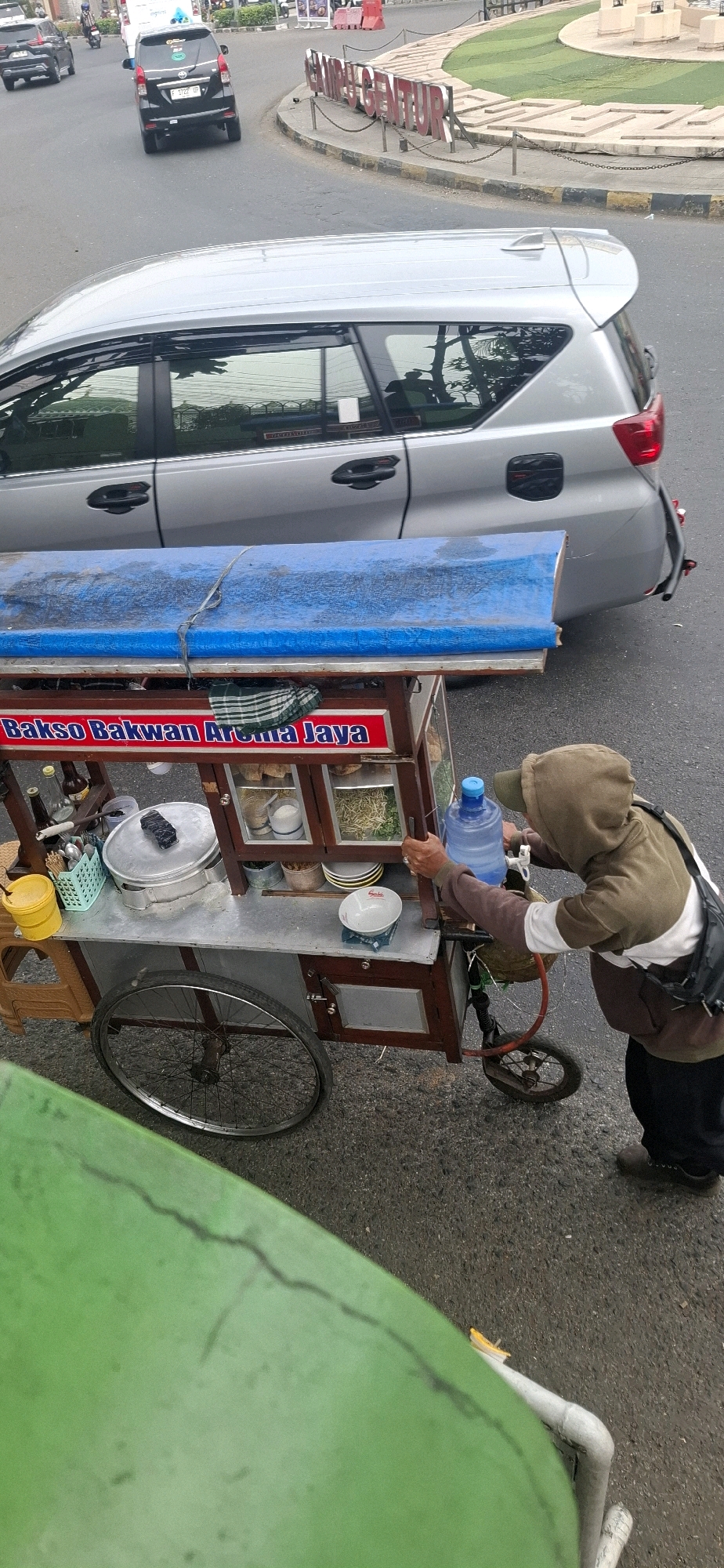 In Cianjur, Indonesia, a street food vendor, wearing a hooded jacket and a cap, is seen tending to their cart. The cart is equipped with a large water dispenser and displays various food items in glass compartments. The cart is situated on an asphalt road, likely in a busy urban area, given the presence of vehicles and a distinctive roadside sign with red lettering that reads "CIBEBER GENTUR". A silver SUV is parked behind the cart, and another car is visible further down the road. The overall scene depicts a typical street commerce setting in the city.