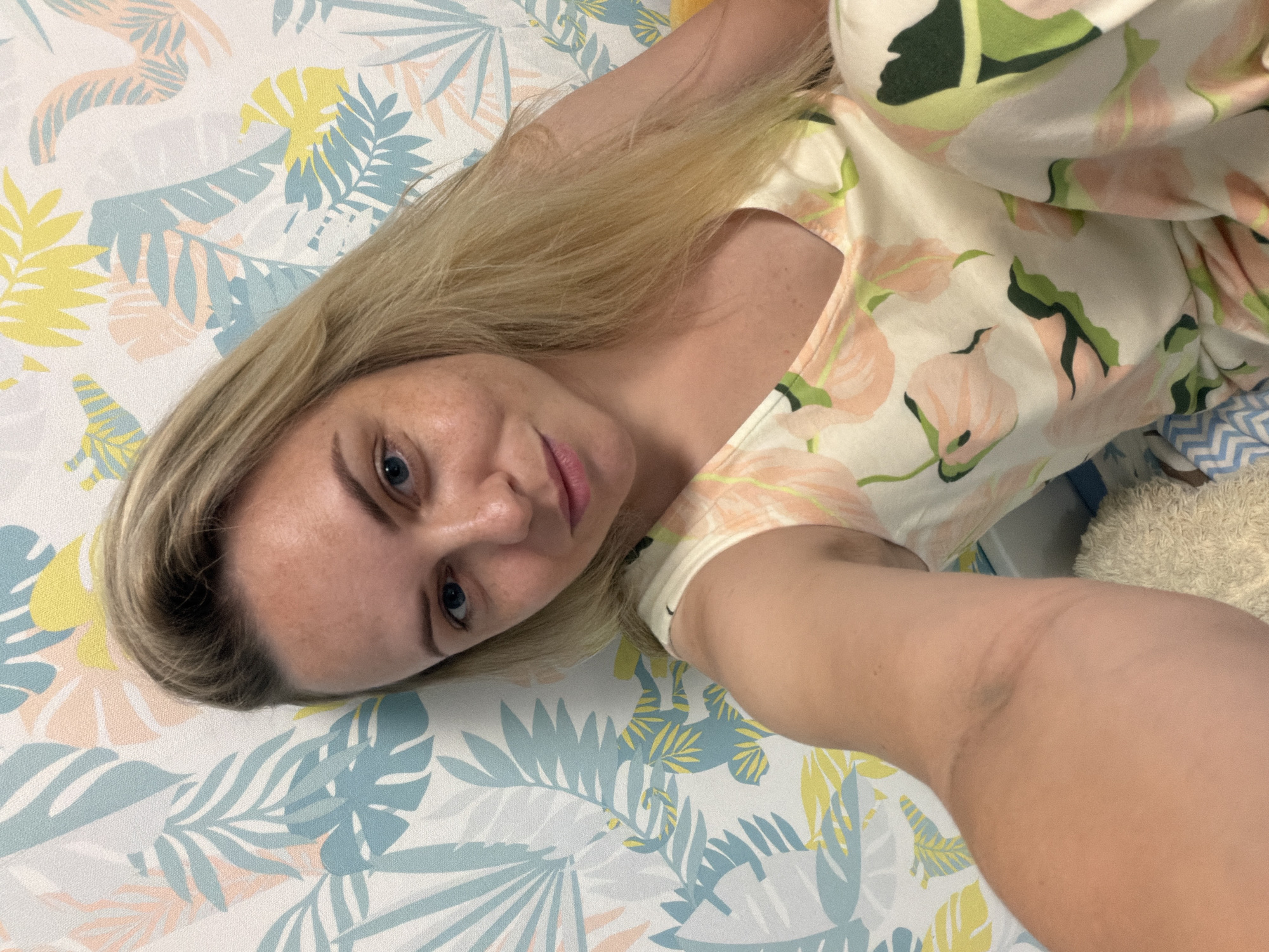 A fair-skinned woman with blonde hair and blue eyes is taking a selfie in front of a patterned wallpaper. The wallpaper features a tropical leaf design in pastel colors of blue, pink, and yellow. The woman is wearing a light-colored dress with a floral pattern of pink and green. Her left arm is extended, holding the camera. The scene appears to be indoors, likely in a bedroom, with hints of a soft toy and bedding visible. The image does not provide specific information about Votkinsk, Russia.