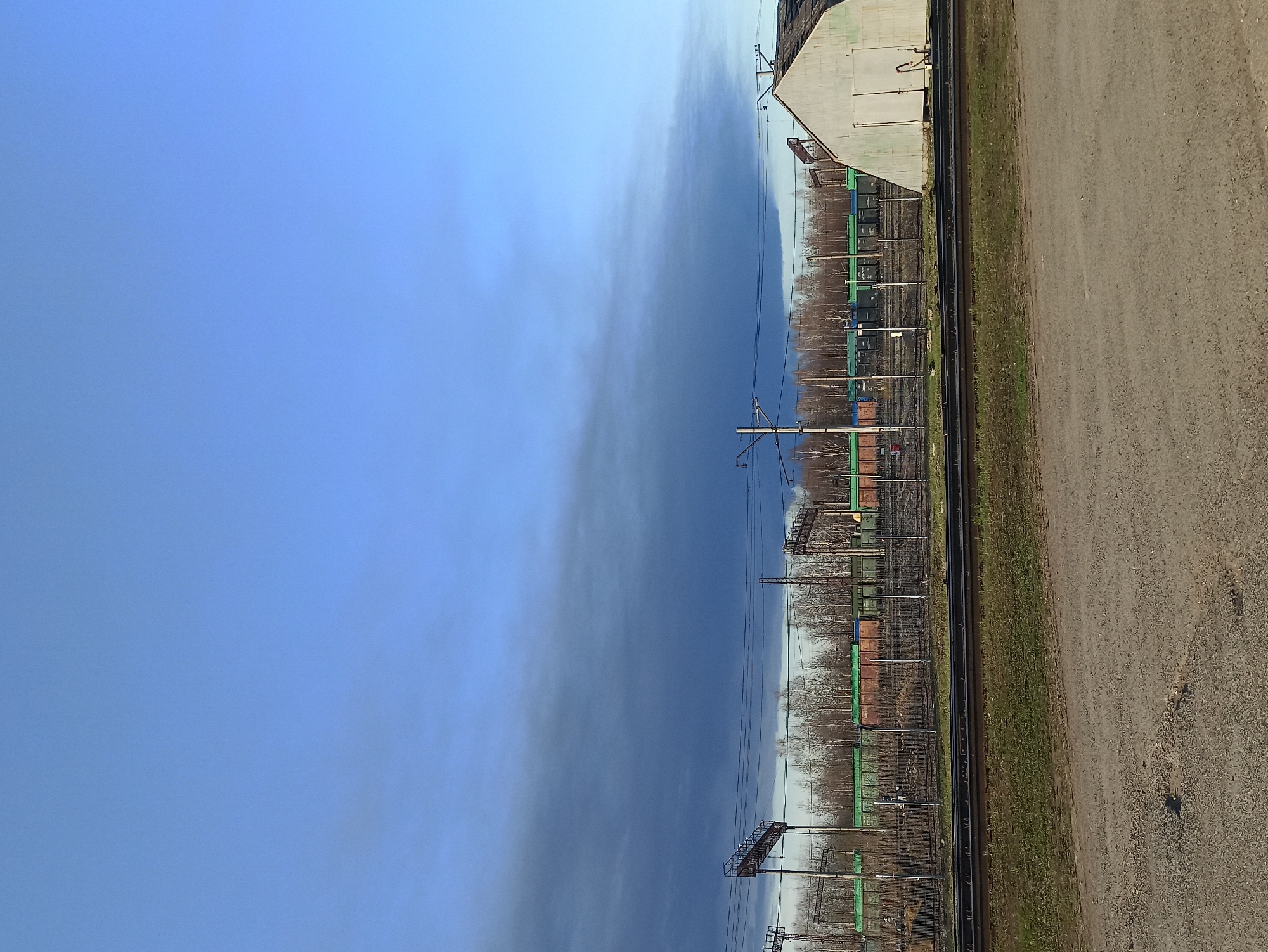 The image displays a landscape featuring a railway and its surroundings under a dynamic sky. The sky, occupying the upper half of the frame, transitions from a bright, clear blue on the left to a darker, hazy or cloudy blue-gray on the right.
Multiple parallel train tracks run horizontally across the middle of the image, receding into the distance. Several train cars, some appearing green and others brownish-red, are present on different tracks, suggesting a freight yard or industrial siding. Alongside the tracks, numerous metal utility poles and overhead line structures, typical for electrified railways, are visible.
Behind the railway lines, a dense row of trees, mostly bare, indicates a setting during autumn, winter, or early spring. To the right, a light-colored, industrial-style building with a sloped roof and several access points or doors is partially visible, situated near the tracks.
The foreground on the right consists of a rough, gravelly or dirt surface, possibly a service road or pathway, with a strip of sparse green grass bordering the railway tracks.
The scene depicts an ordinary daytime view of railway infrastructure. There are no people visible, nor any specific activities taking place. No discernible text is present in the image.