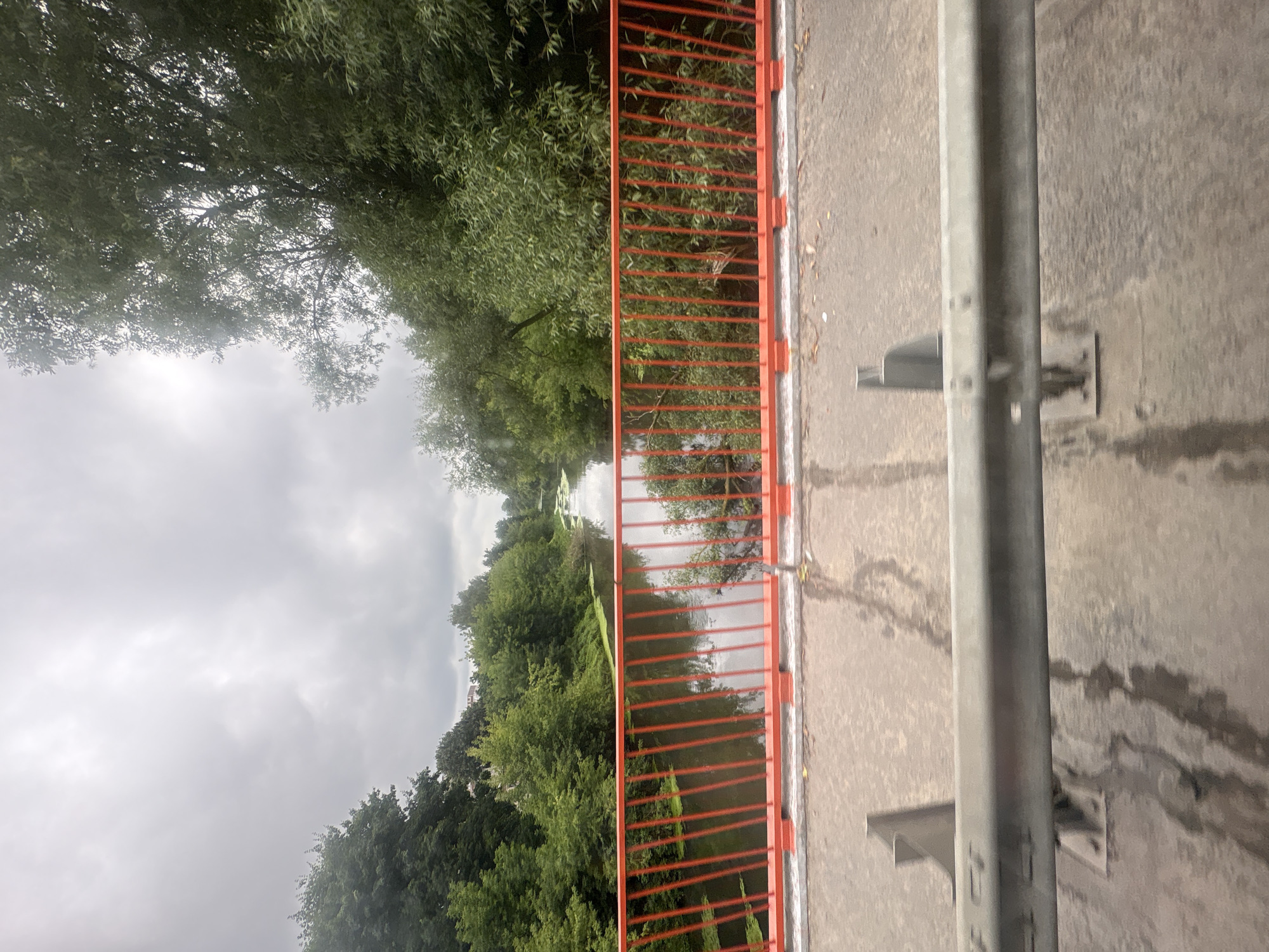 This is a ground-level, eye-level, medium shot of a river with lush green trees on both banks. The river is somewhat narrow and appears to be partially covered with green algae or duckweed. A bright orange metal railing, a common feature of pedestrian bridges or overpasses, runs horizontally across the foreground, obscuring the lower portion of the scene. The railing is made of vertical bars and a horizontal top rail. The ground in the foreground appears to be asphalt, with some visible reflections, suggesting it might be wet. The sky is overcast, with heavy, gray clouds. The scene is likely a bridge overlooking a river in a natural setting within the city of Sverdlovskiy, Russia. There are no people visible in the image.